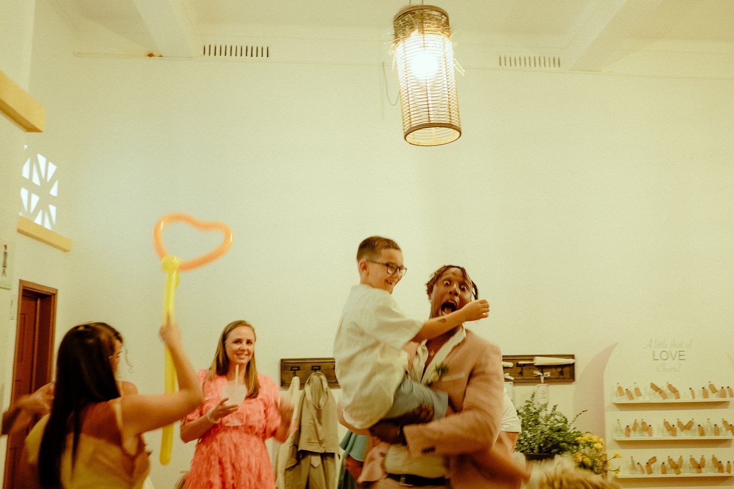 A joyful man lifts a young boy at a wedding celebration while guests smile and hold colorful bubble wands indoors.