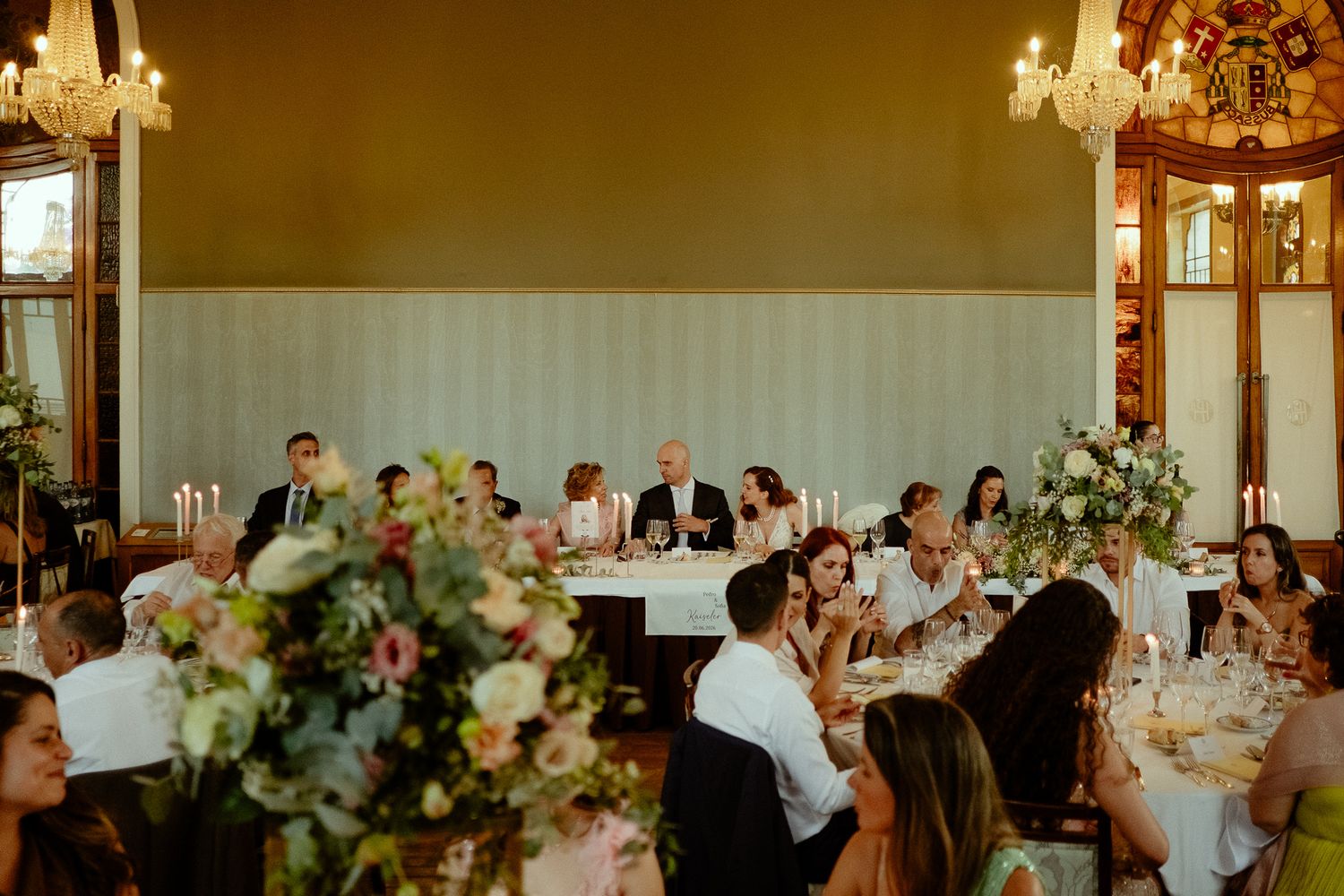 Elegant wedding reception in a grand ballroom with floral centerpieces, candlelight, and guests gathered around tables during speeches.