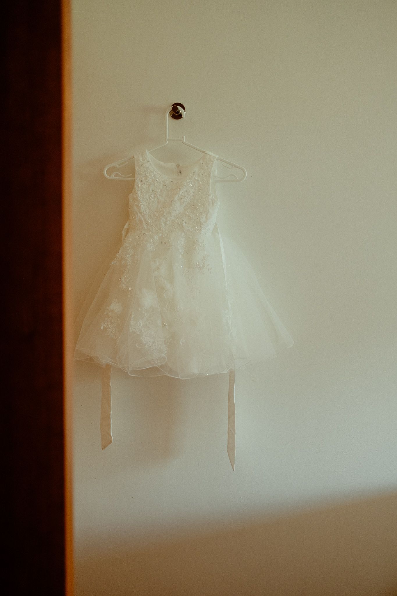 White lace flower girl dress hanging on a hanger against a cream wall, perfect for a wedding ceremony.