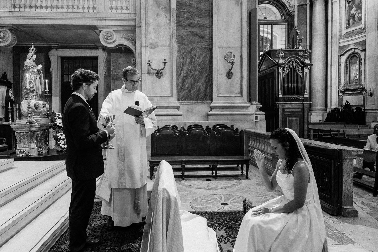 Black and white wedding ceremony photo inside ornate church with bride, groom, and officiant at the altar.