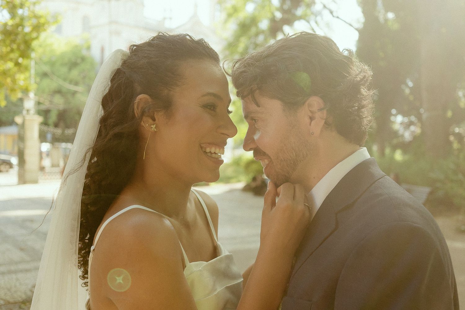 A smiling bride and groom share an intimate moment outdoors on their wedding day in warm sunlight.