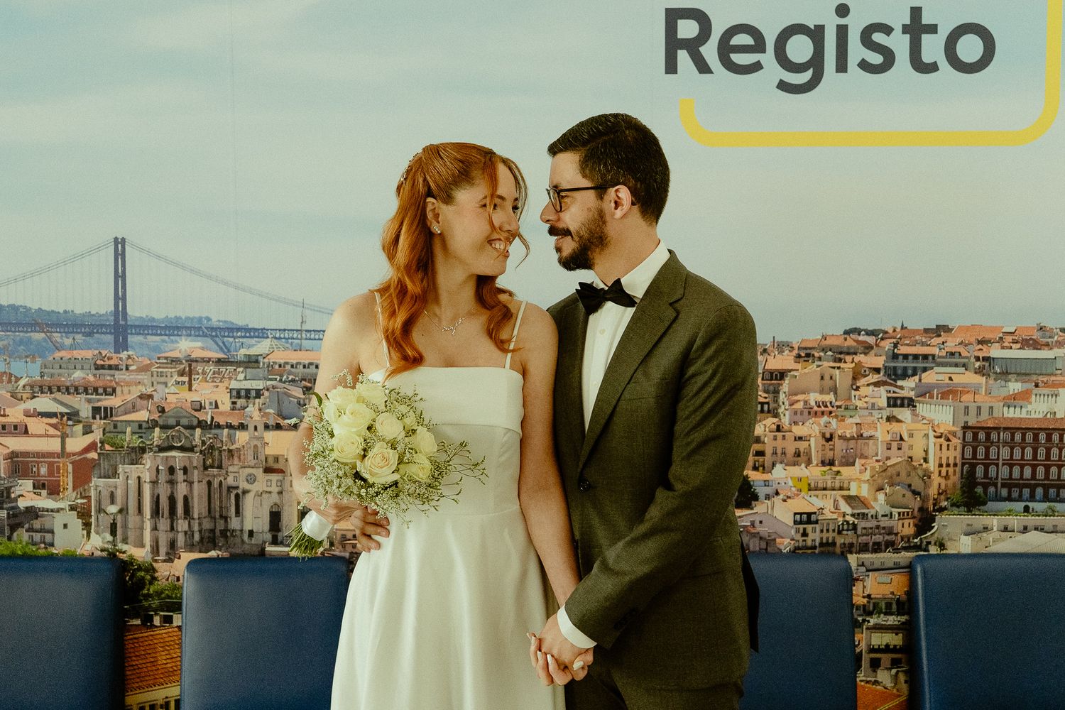 A bride and groom hold hands smiling at each other with a Lisbon cityscape and bridge backdrop.