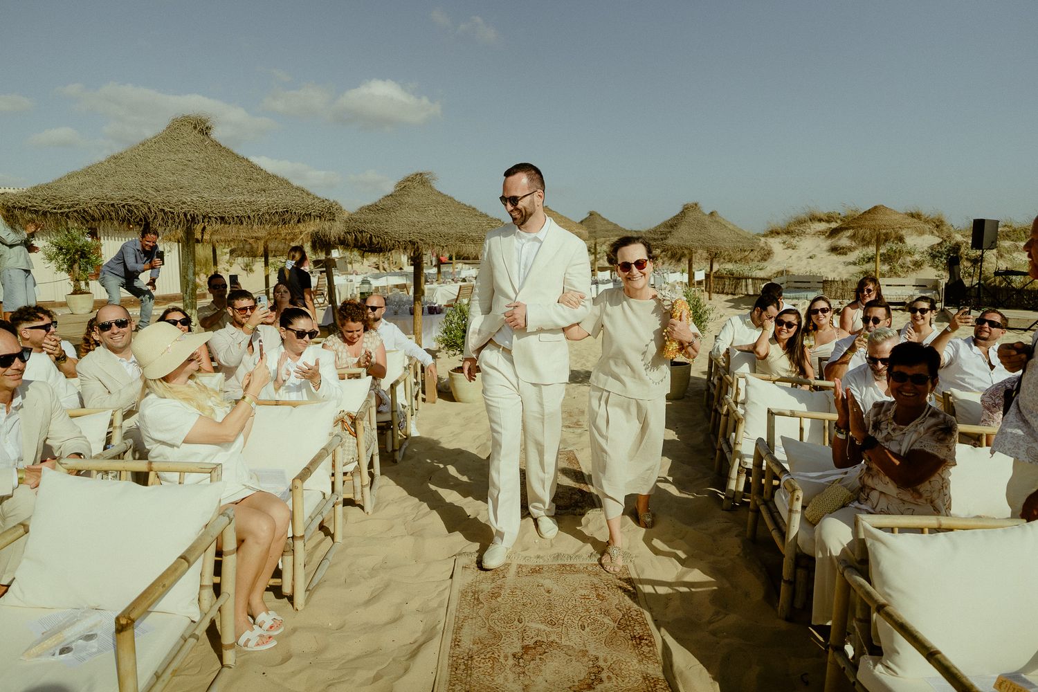 Two grooms in white suits walk through cheering guests at a beach wedding ceremony with thatched umbrellas.