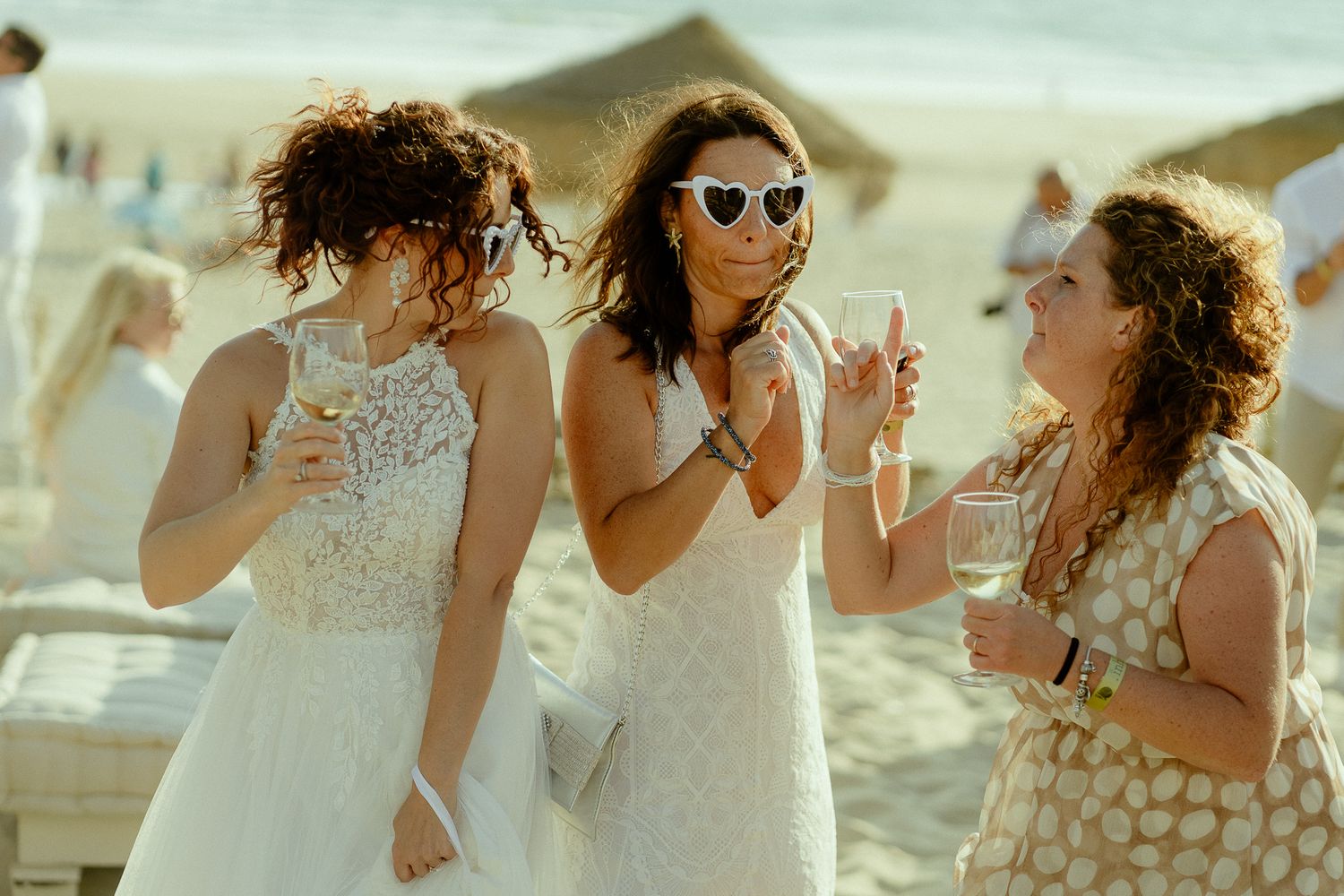 Three women in white dresses socializing on a sunny beach, holding drinks and smiling at an outdoor event.