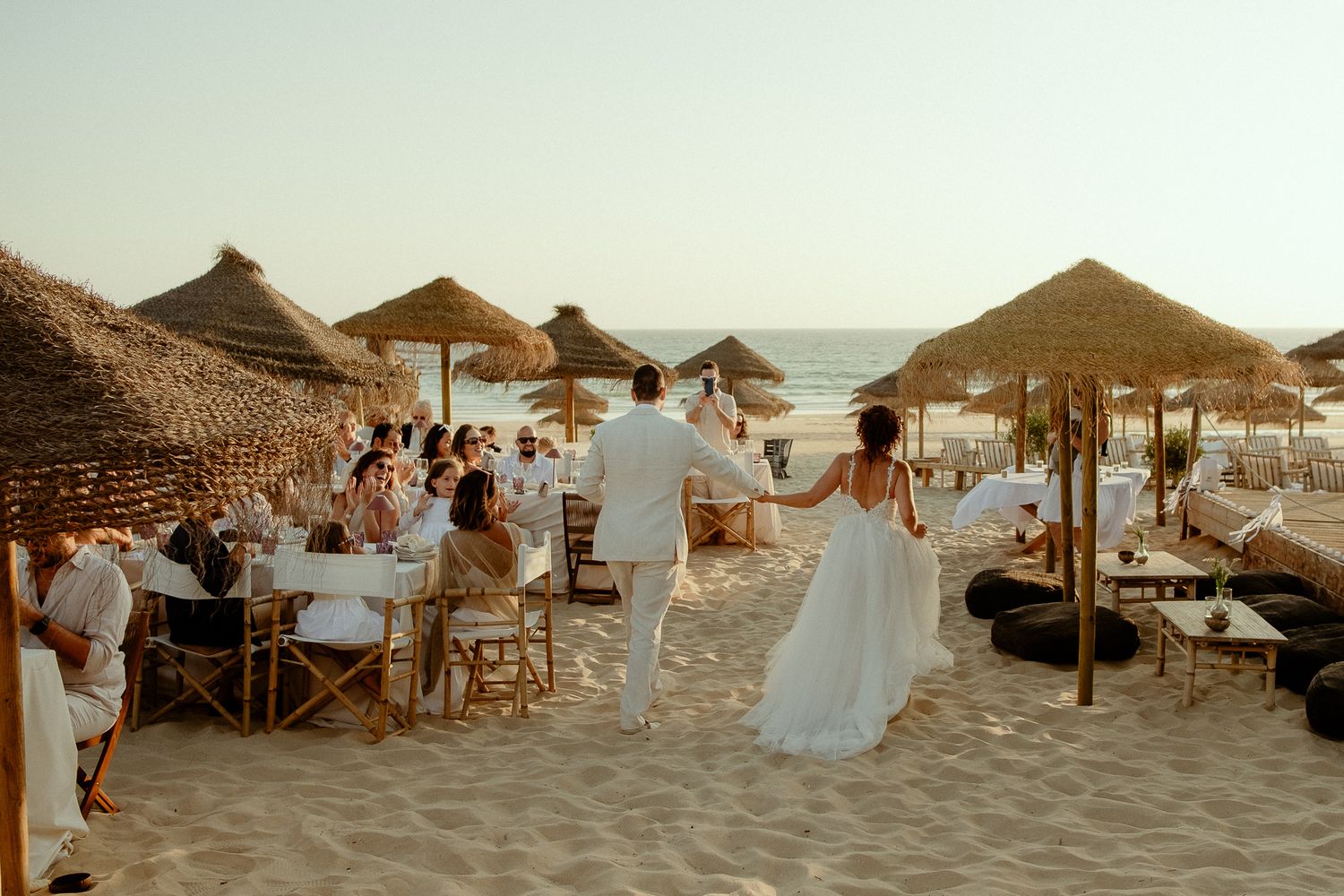 Bride and groom walk hand-in-hand on a sandy beach wedding reception with straw umbrellas and guests seated.