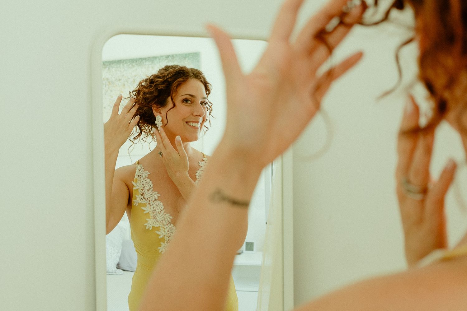 Bride admiring herself in a mirror while putting on earrings, wearing a yellow floral dress before her wedding.