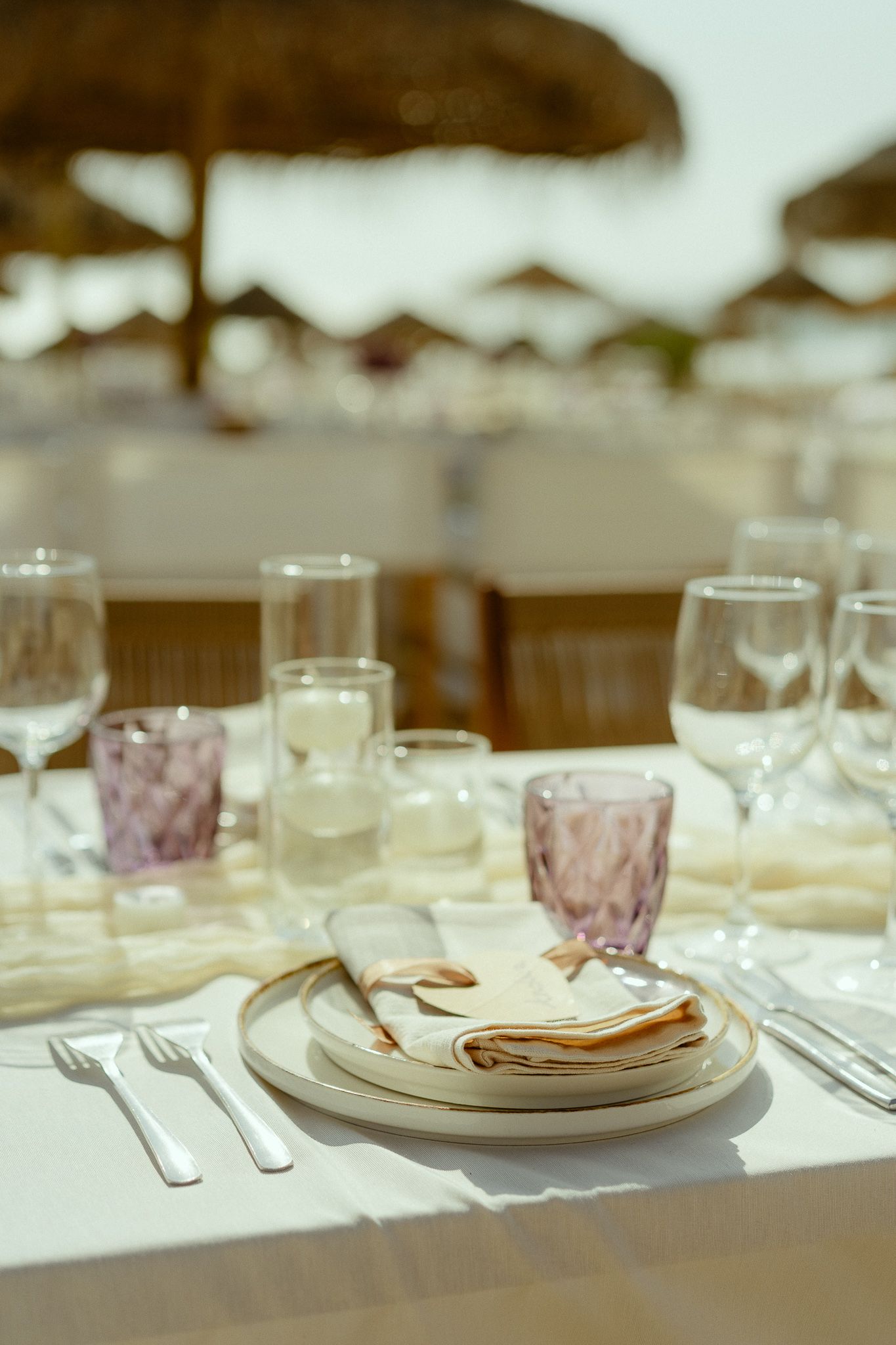 Elegant beach restaurant table setting with white plates, purple glasses, wine glasses, and silverware under thatched umbrellas.