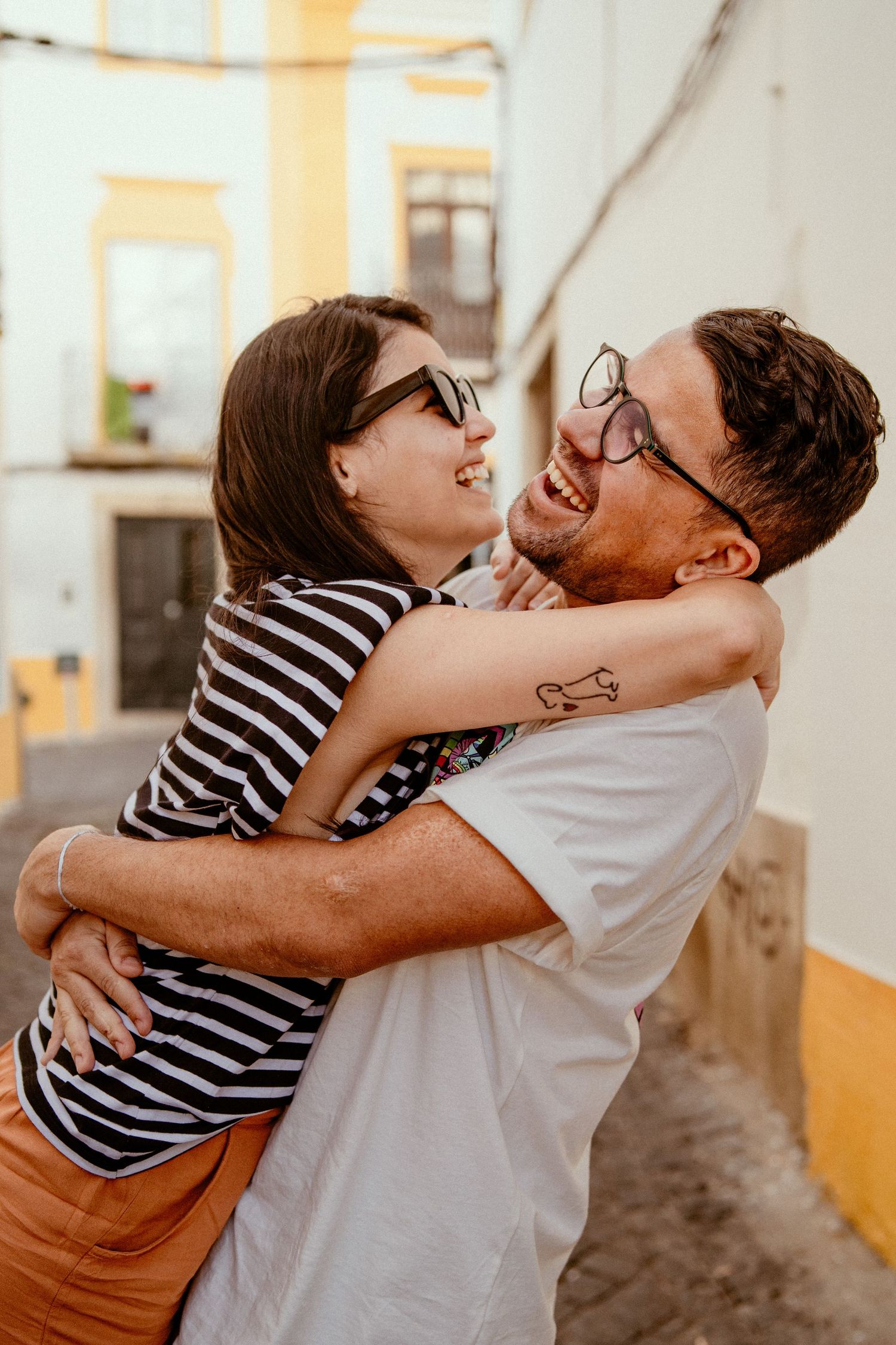 Happy couple wearing glasses embrace and laugh together on a sunny street with yellow and white walls.