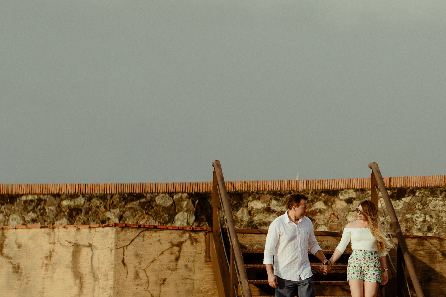 A couple walks hand in hand through a desert landscape at sunset with rock formations in the background.