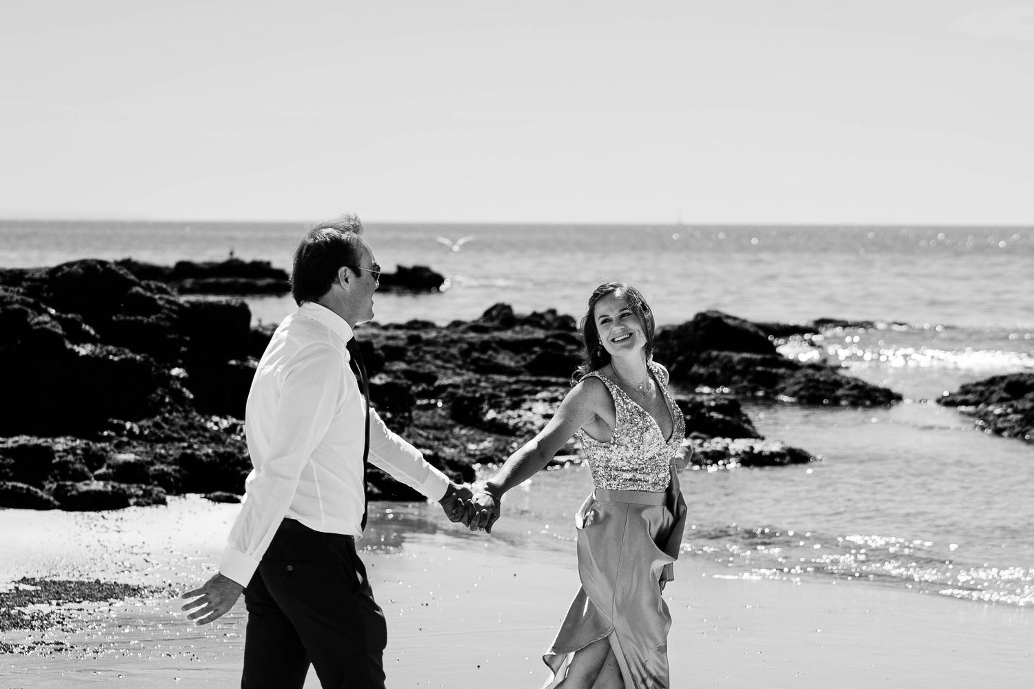 A couple holds hands while walking along a rocky beach shoreline in black and white photography.