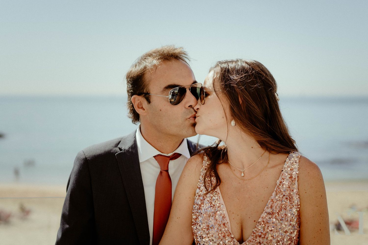 A romantic couple shares a tender kiss by the ocean with the groom in a suit and bride in a sparkly dress.