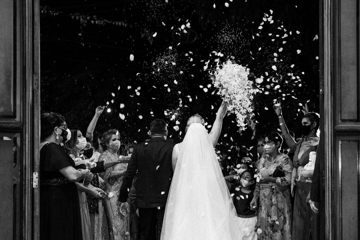 Wedding guests shower newlyweds with confetti at church doorway in dramatic black and white photograph.