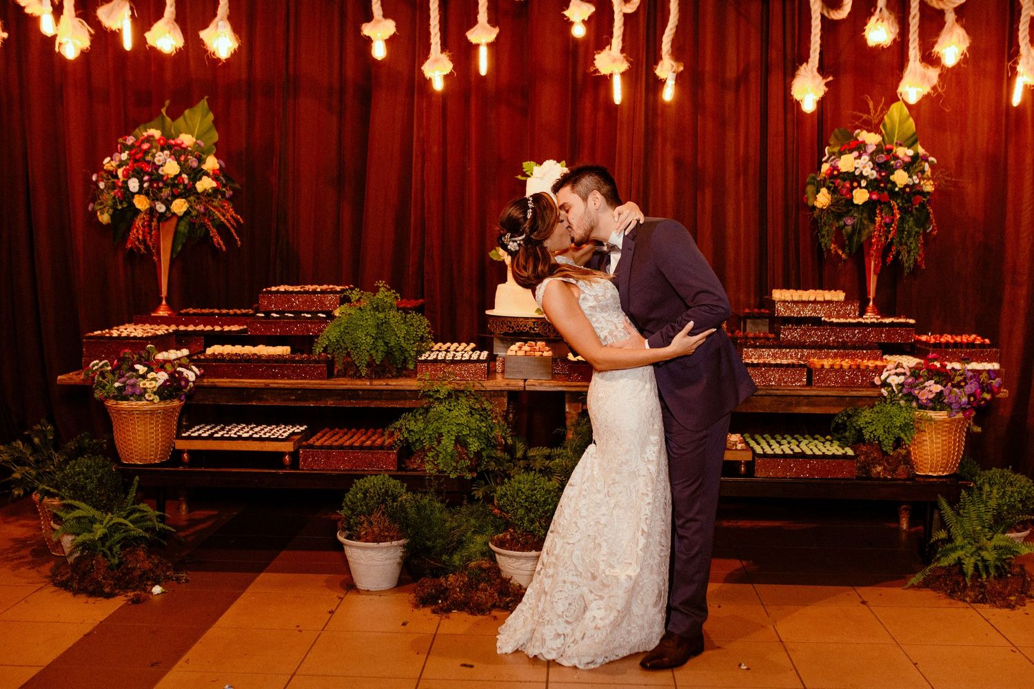 A romantic couple shares their first dance on a dimly lit dance floor with wooden paneled walls and potted plants.