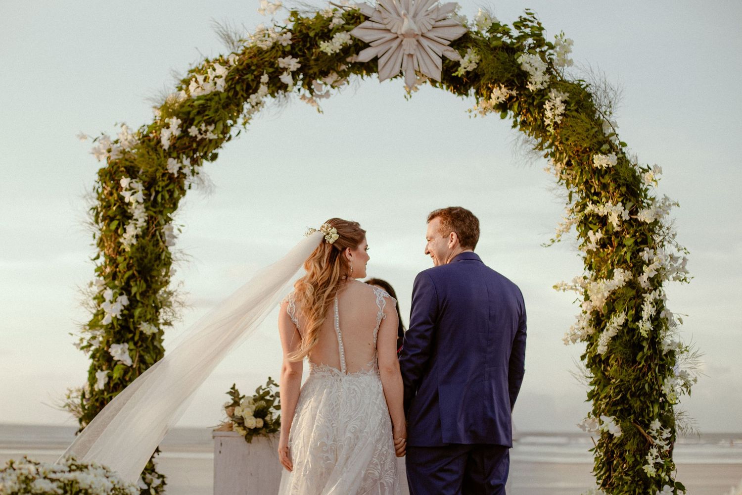 A wedding couple stands under a floral arch decorated with greenery and lights against a sunset sky.
