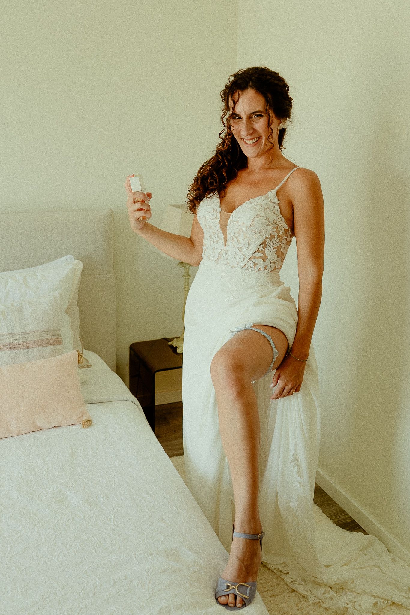 Smiling bride in a white lace wedding gown with a thigh-high slit, wearing a garter and holding a perfume bottle.