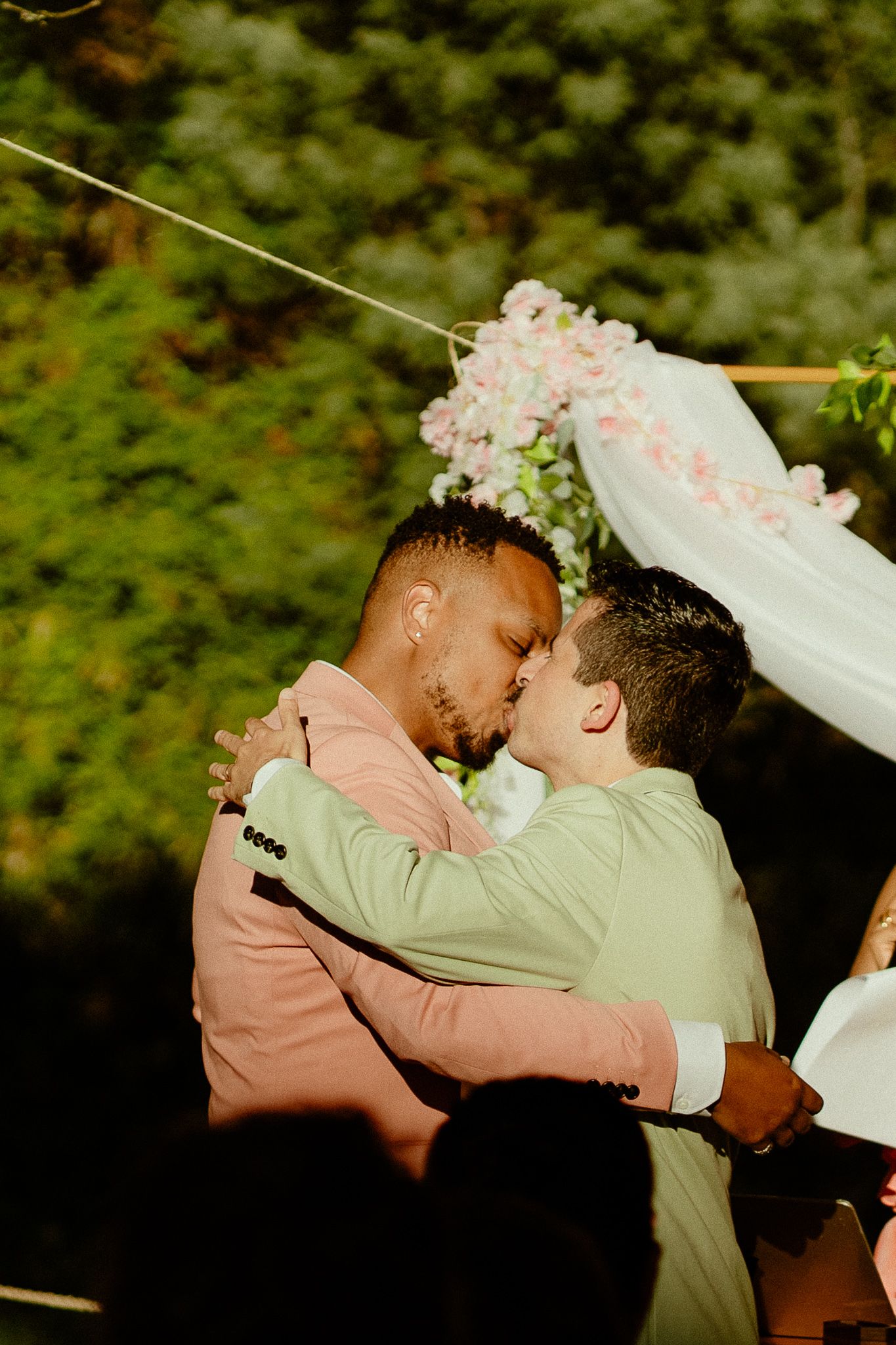 Two grooms share a first kiss at outdoor wedding ceremony beneath floral arch with pink blooms and white draping.
