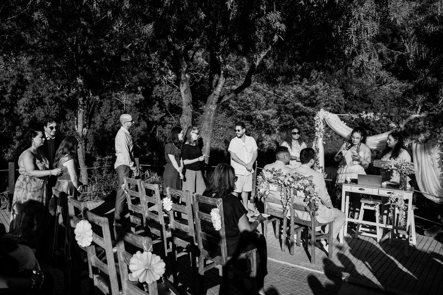 Black and white outdoor wedding ceremony with guests gathered among trees on a wooden deck.