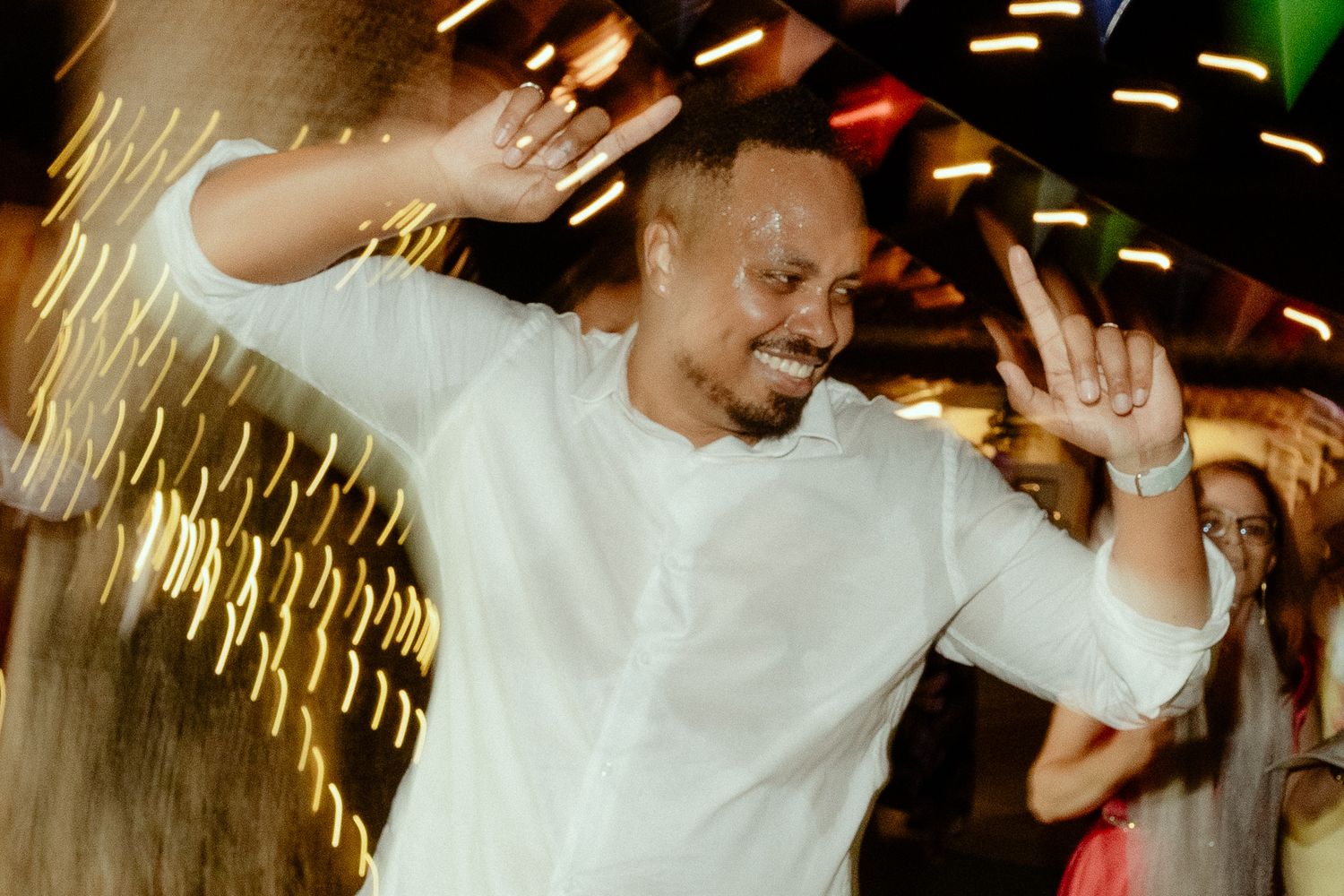 Man in white shirt dancing joyfully at a party with colorful bokeh lights in the background.