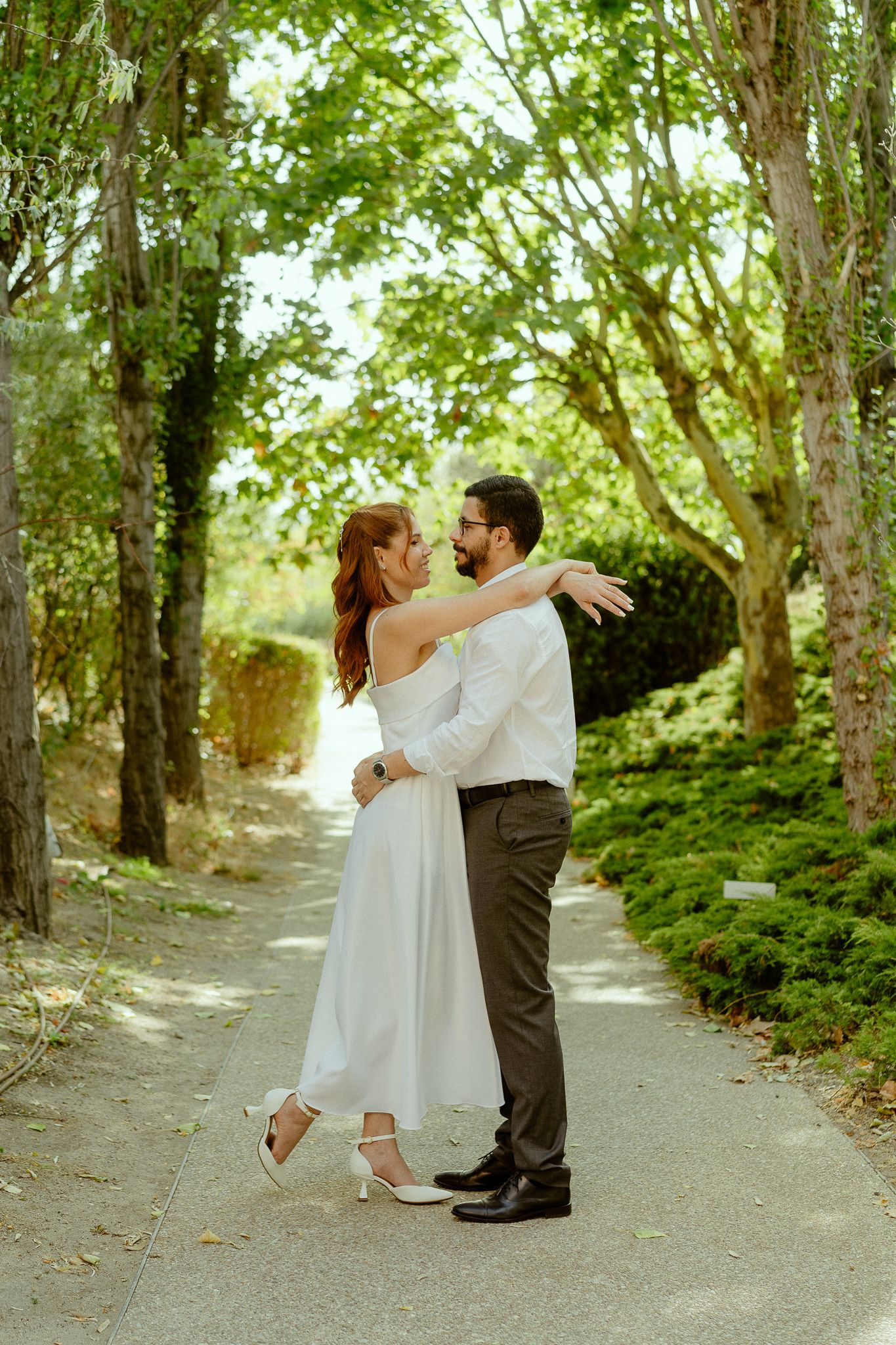 Bride in white dress and groom in suit embrace on a tree-lined garden path on their wedding day.