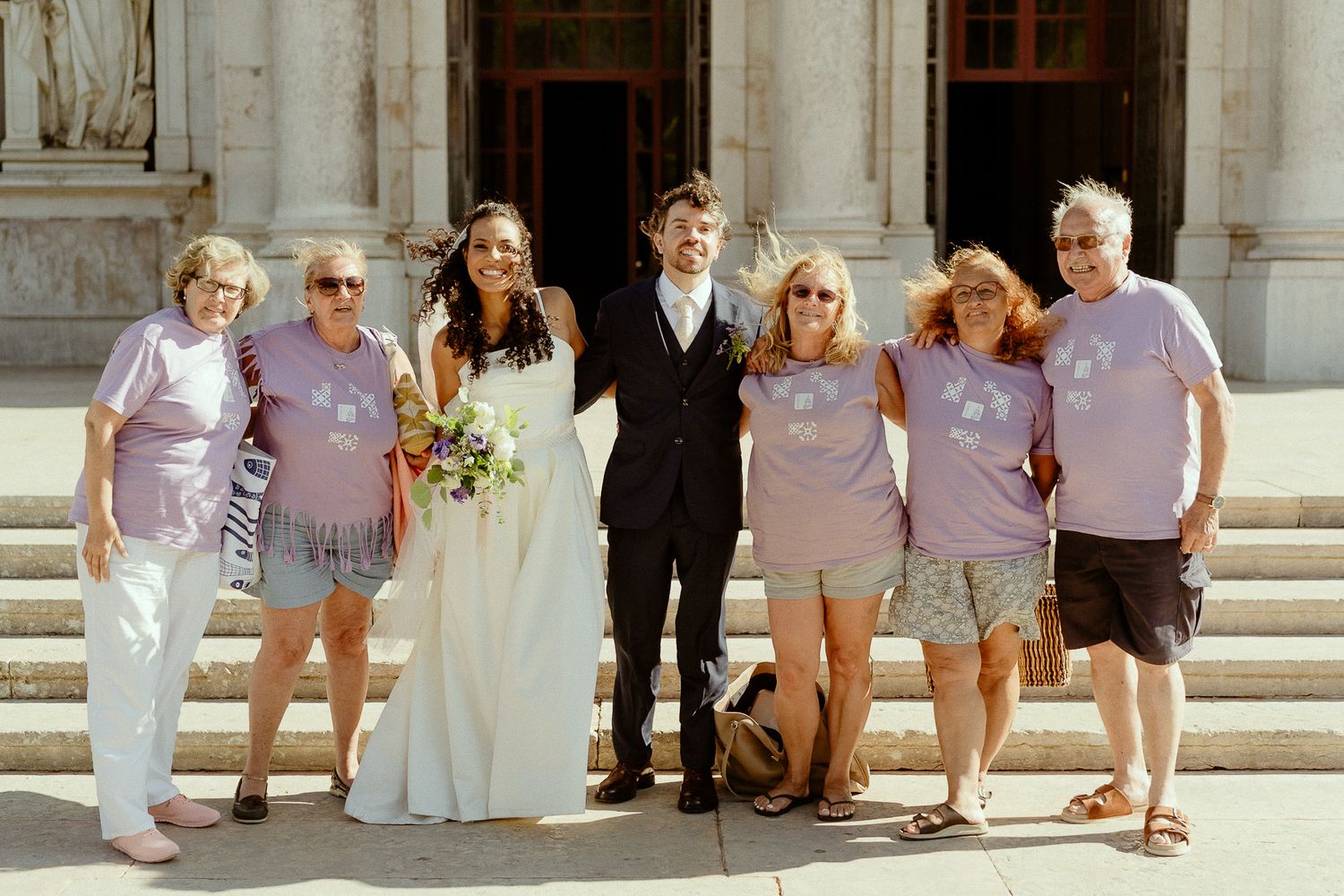 Wedding couple in white poses with family members wearing matching pink shirts outside a grand stone building.