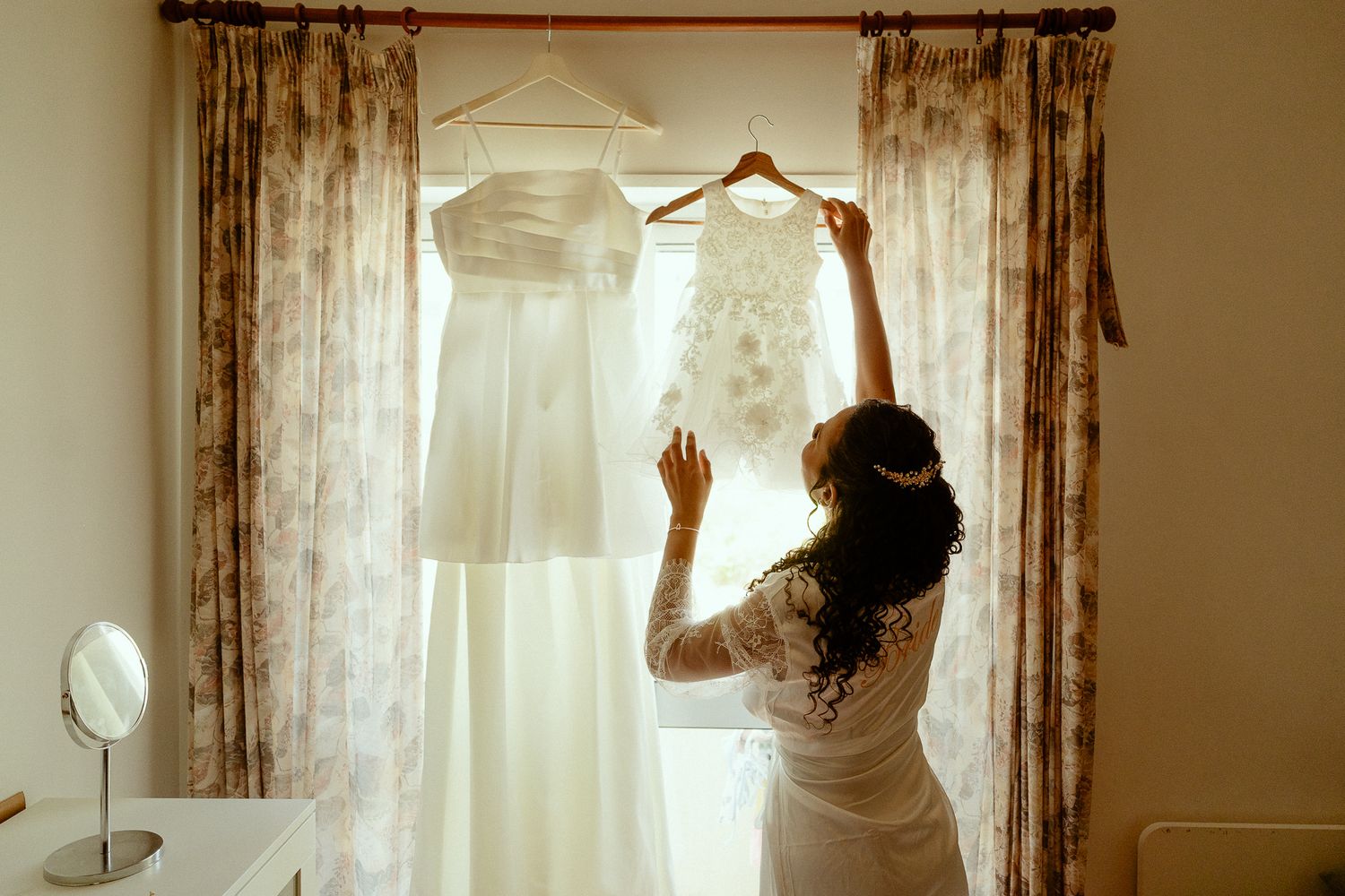 Bride in white lace robe holding wedding dress on hanger near curtained window with mirror on dresser.