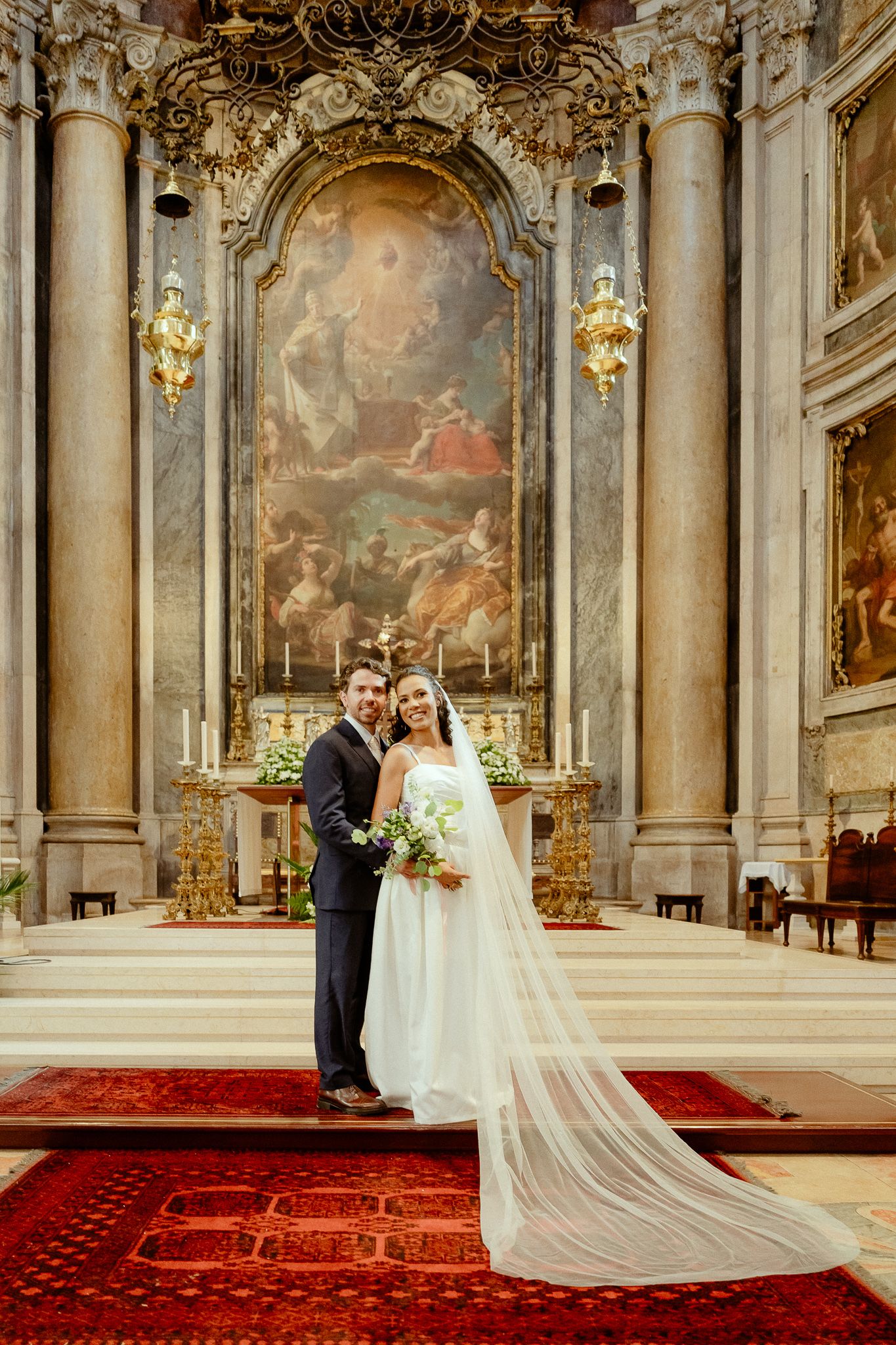 Bride and groom pose at ornate baroque church altar with gold details and classical painting backdrop.