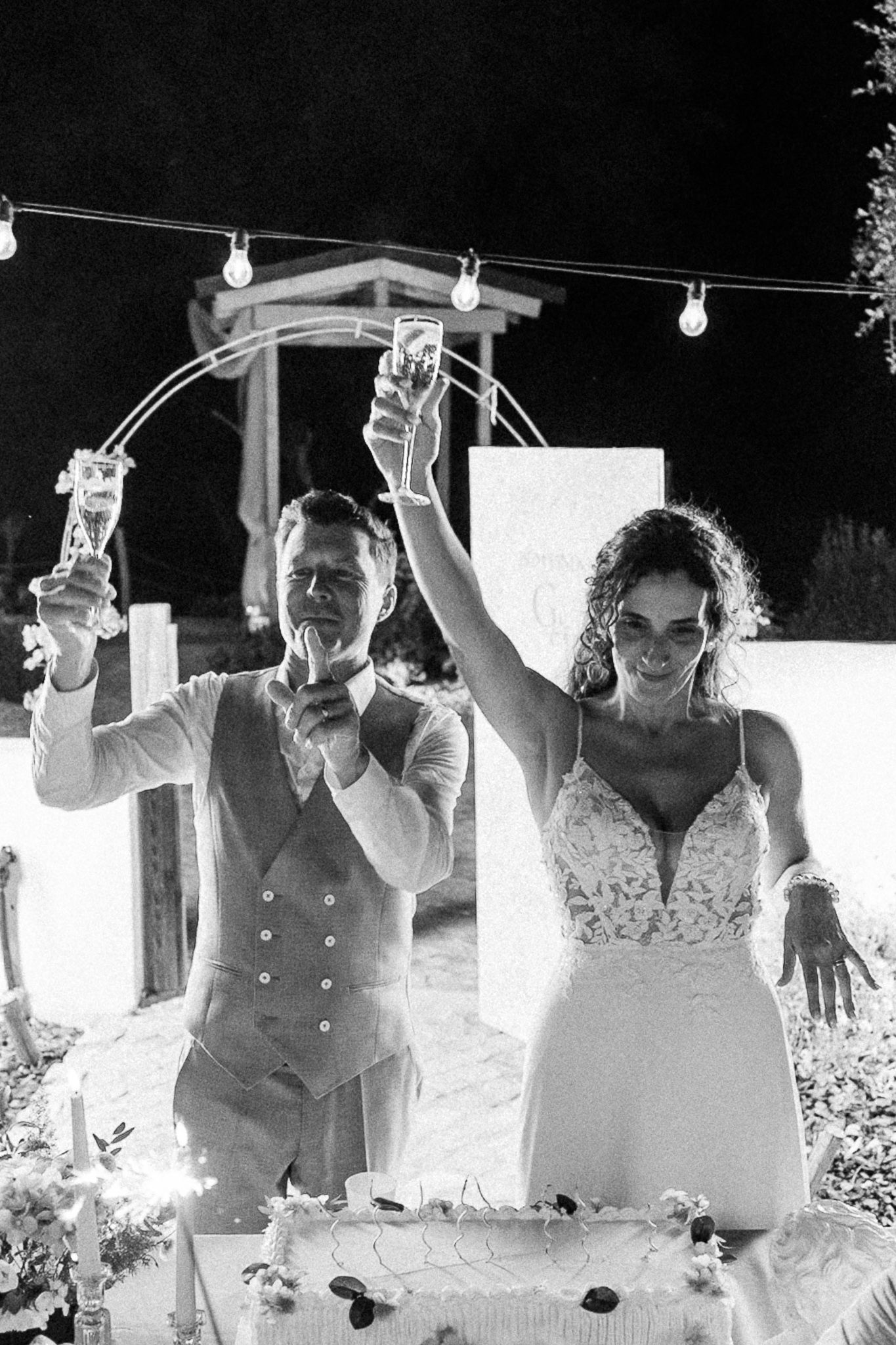 Black and white photo of bride and groom raising champagne glasses over wedding cake at outdoor night reception.