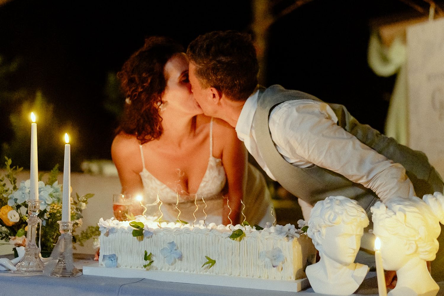 Newlywed couple sharing a romantic kiss over white wedding cake with candles at outdoor night reception.