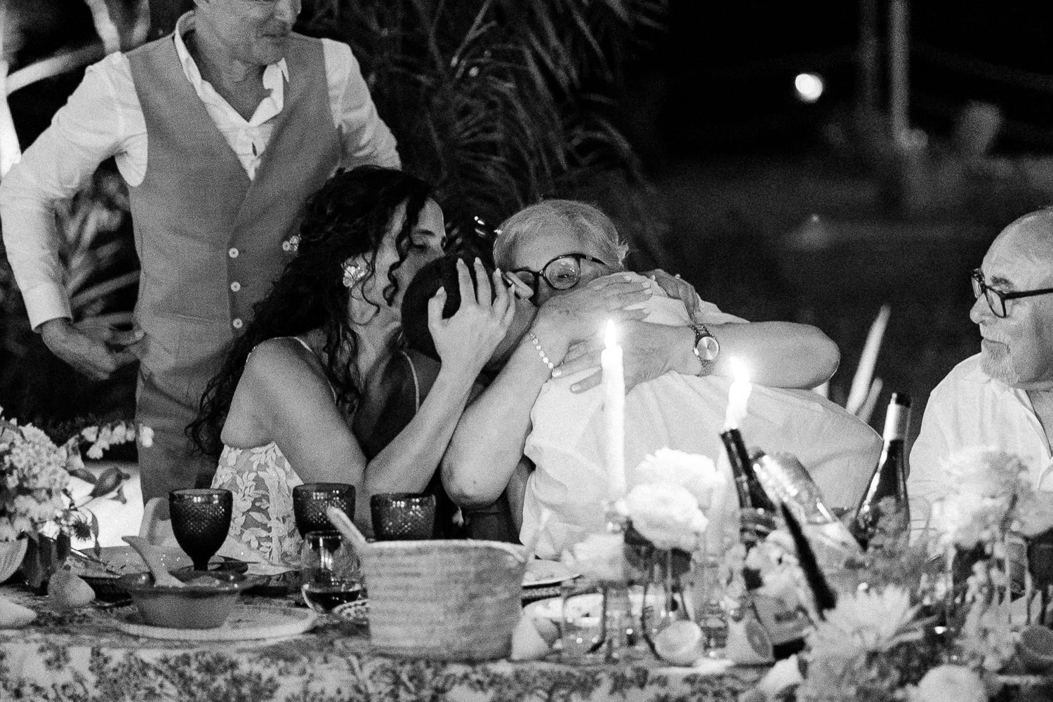 Black and white wedding reception photo of guests embracing at candlelit table with floral arrangements.