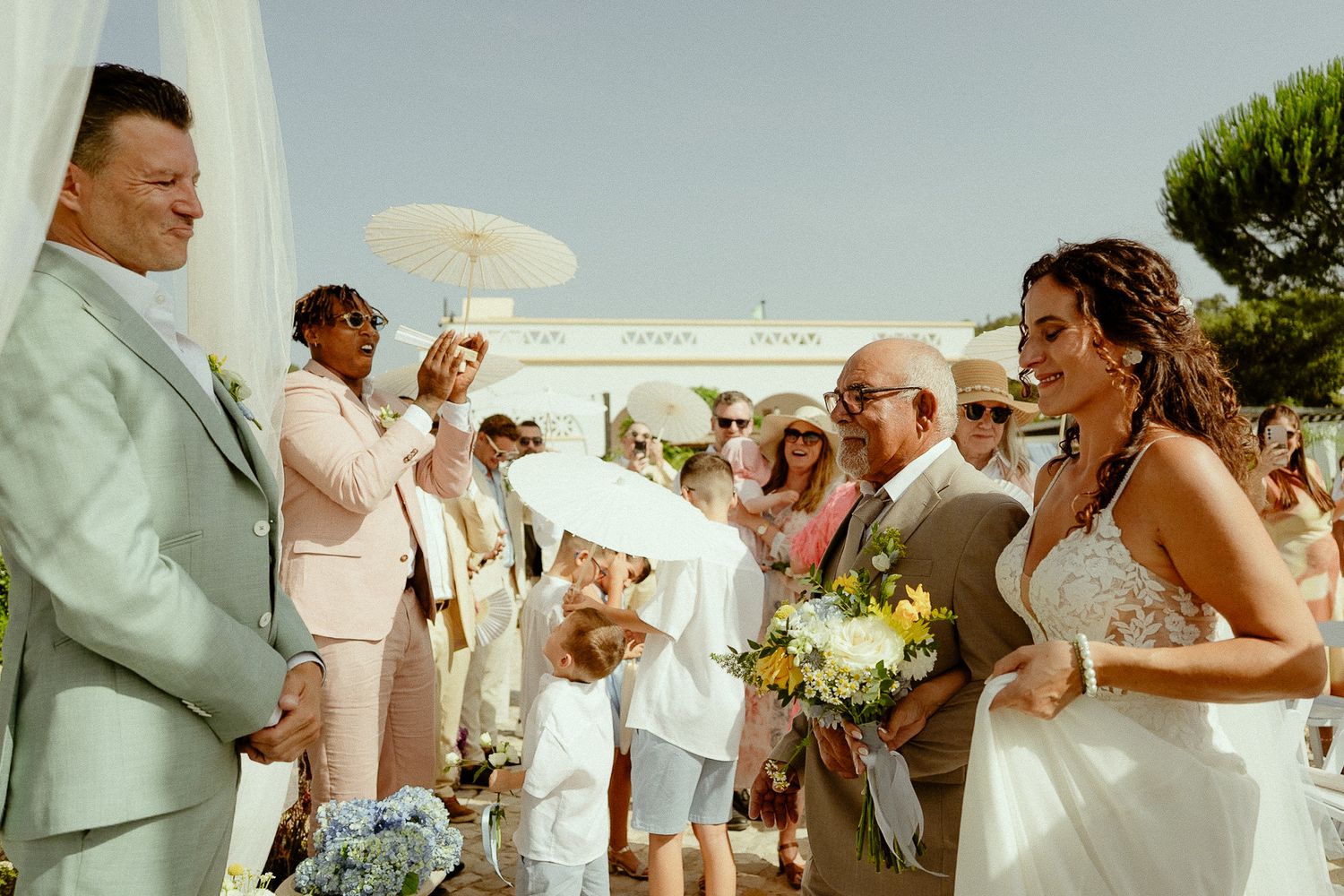 Bride and groom exchange vows outdoors at a sunny wedding ceremony with guests and a flower girl nearby.