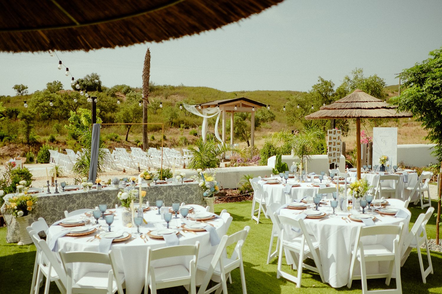 Elegant outdoor wedding reception with white round tables, chairs, and thatched umbrellas set on green grass under sunny skies.
