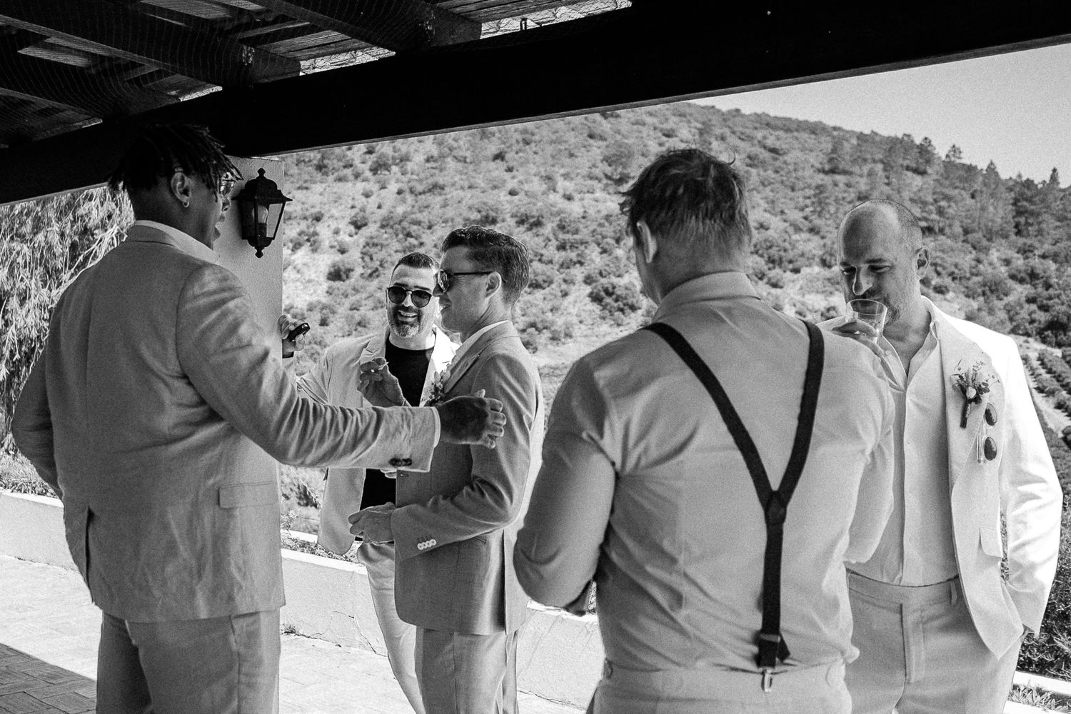 Black and white photo of groomsmen in suits sharing a moment before a wedding ceremony outdoors.