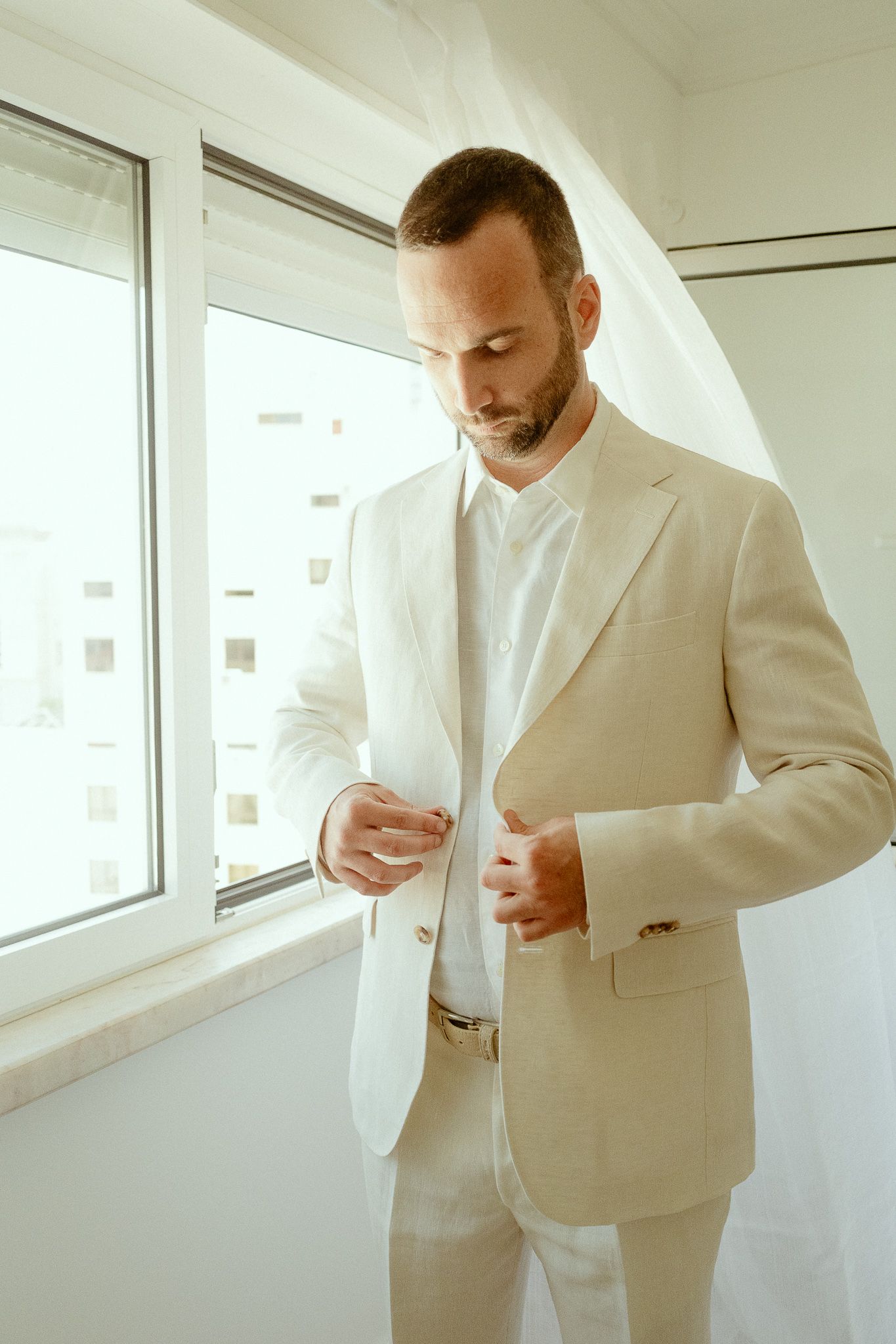 A well-dressed groom in a beige linen suit buttoning his jacket while standing near a bright window.