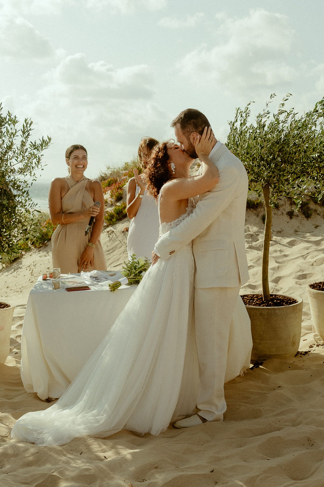 Bride and groom share first kiss on sandy beach wedding, bridesmaid smiling behind, olive trees and table in background.