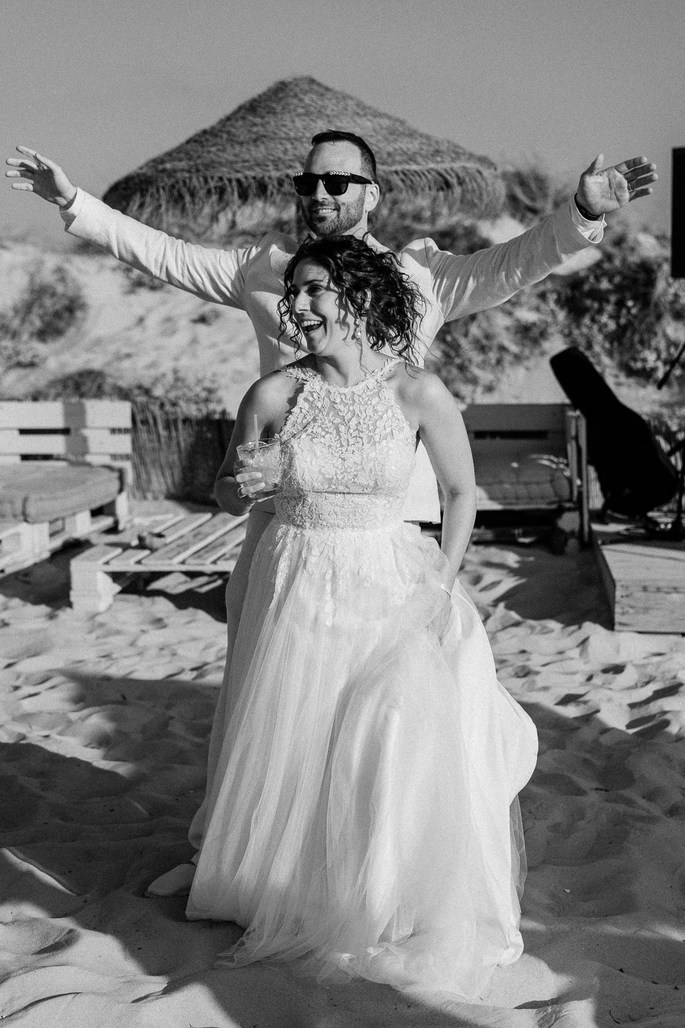 Black and white photo of a joyful bride and groom celebrating on a sandy beach, groom with arms outstretched wide.