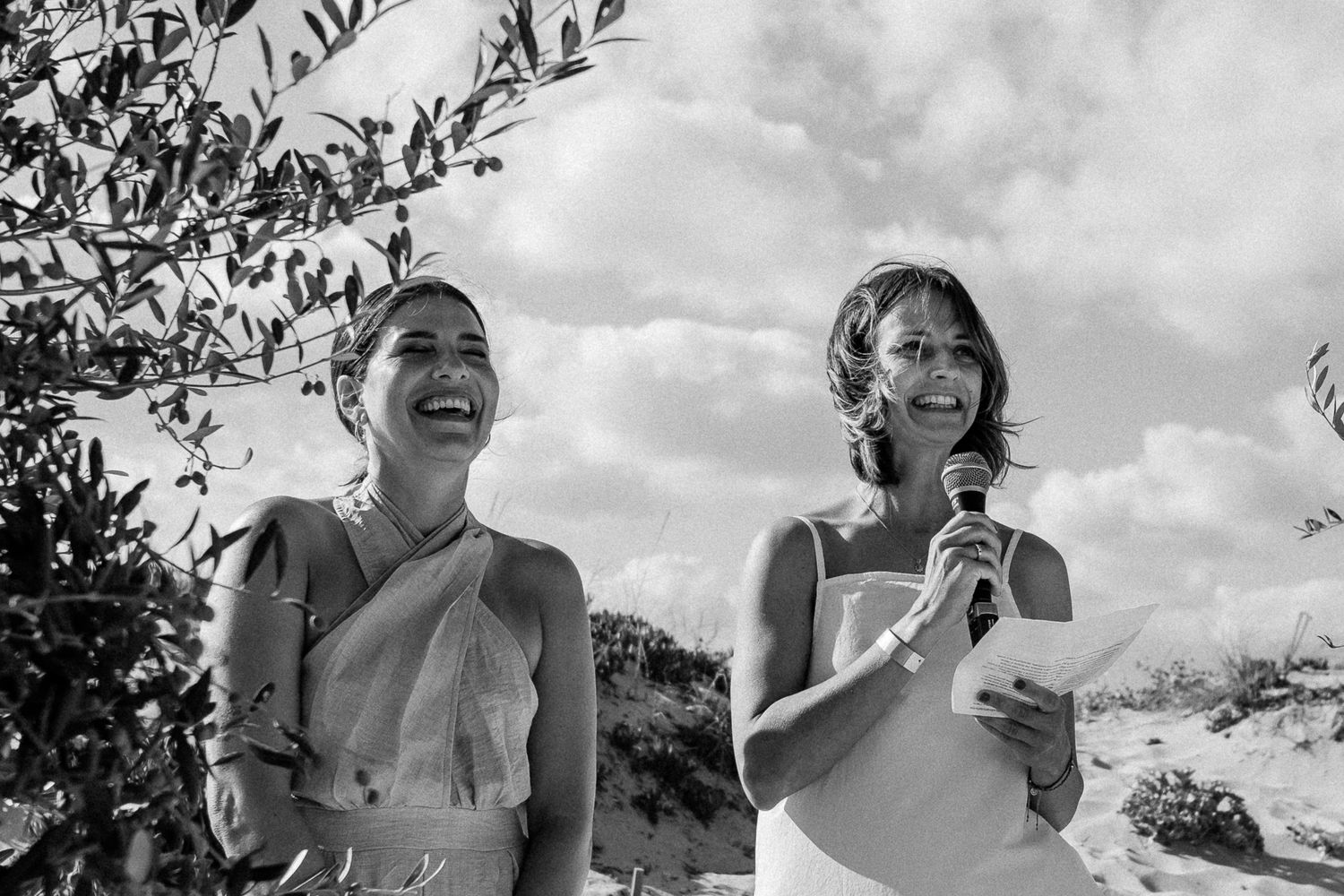 Two women laughing and speaking into a microphone outdoors at a wedding ceremony on a sunny day.