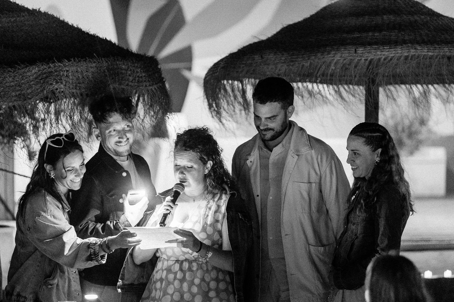 Group of friends celebrating with sparklers outdoors at night near thatched umbrellas in black and white photo.