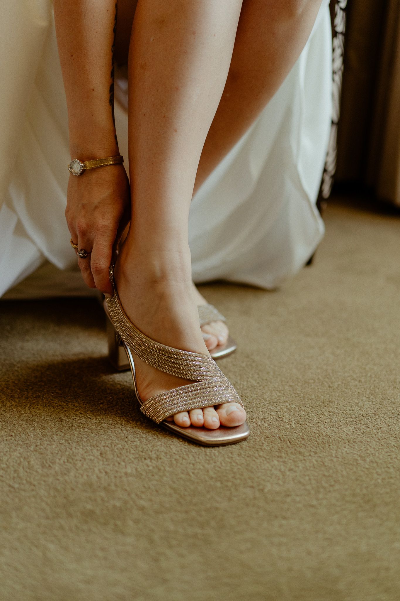 Bride putting on elegant gold strappy heeled sandals while wearing a white wedding dress.