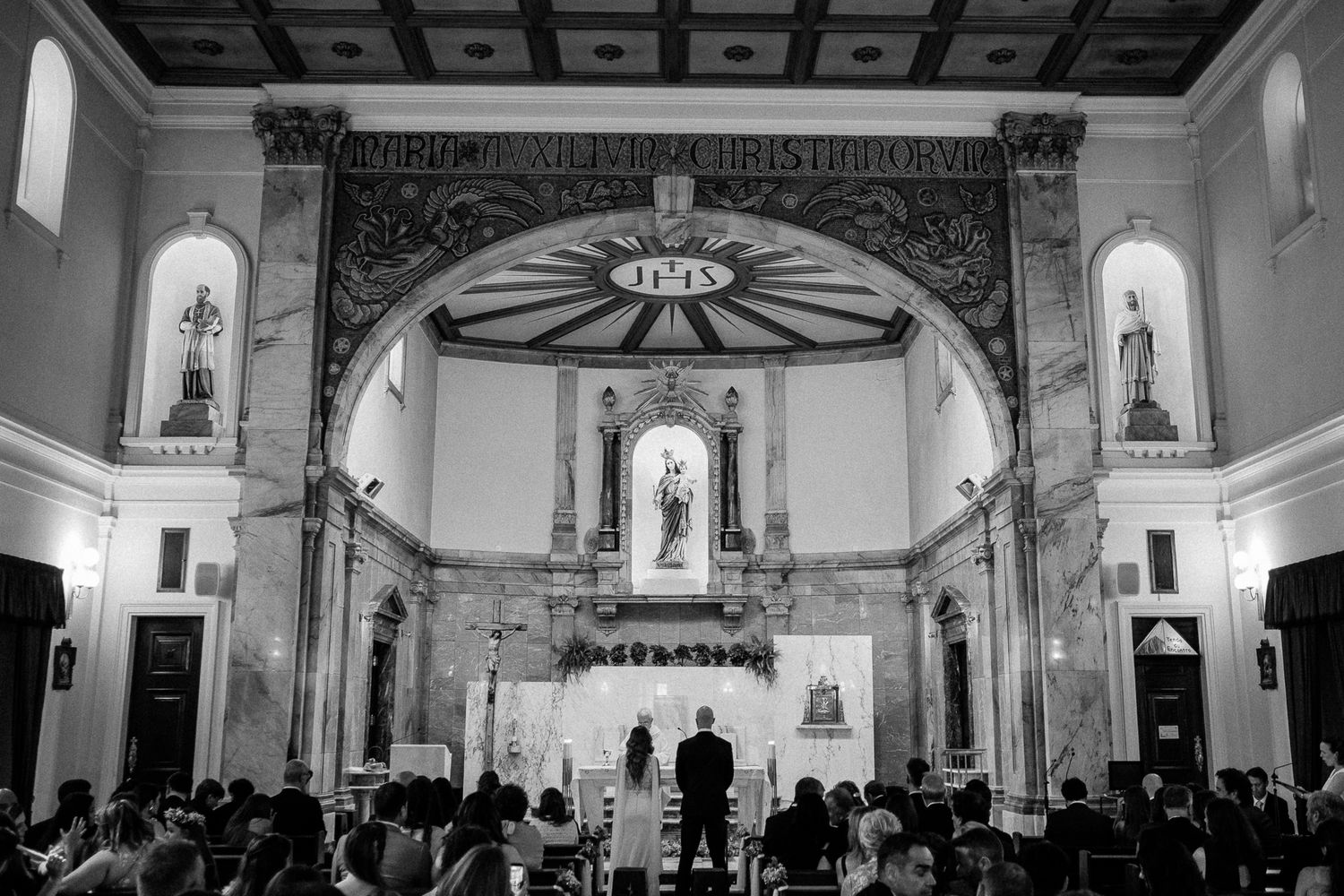 Black and white photo of a wedding ceremony inside an ornate Catholic church with guests seated in pews.