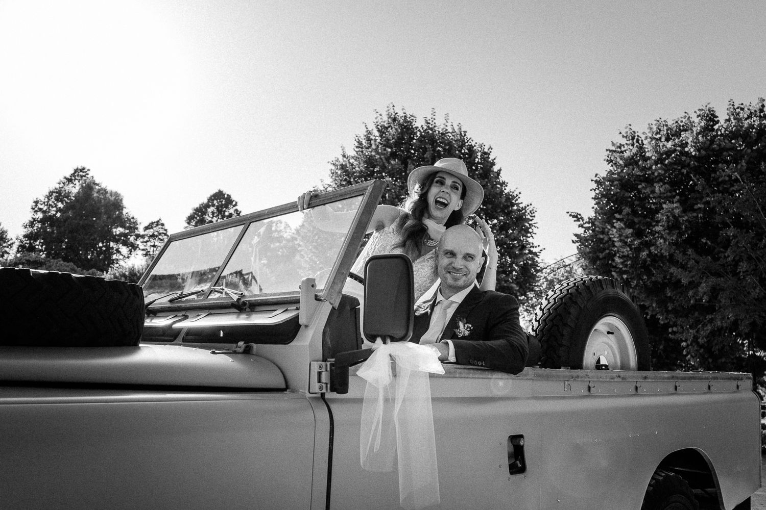 Black and white photo of a smiling couple in a vintage open-top car decorated with white ribbons at a wedding.