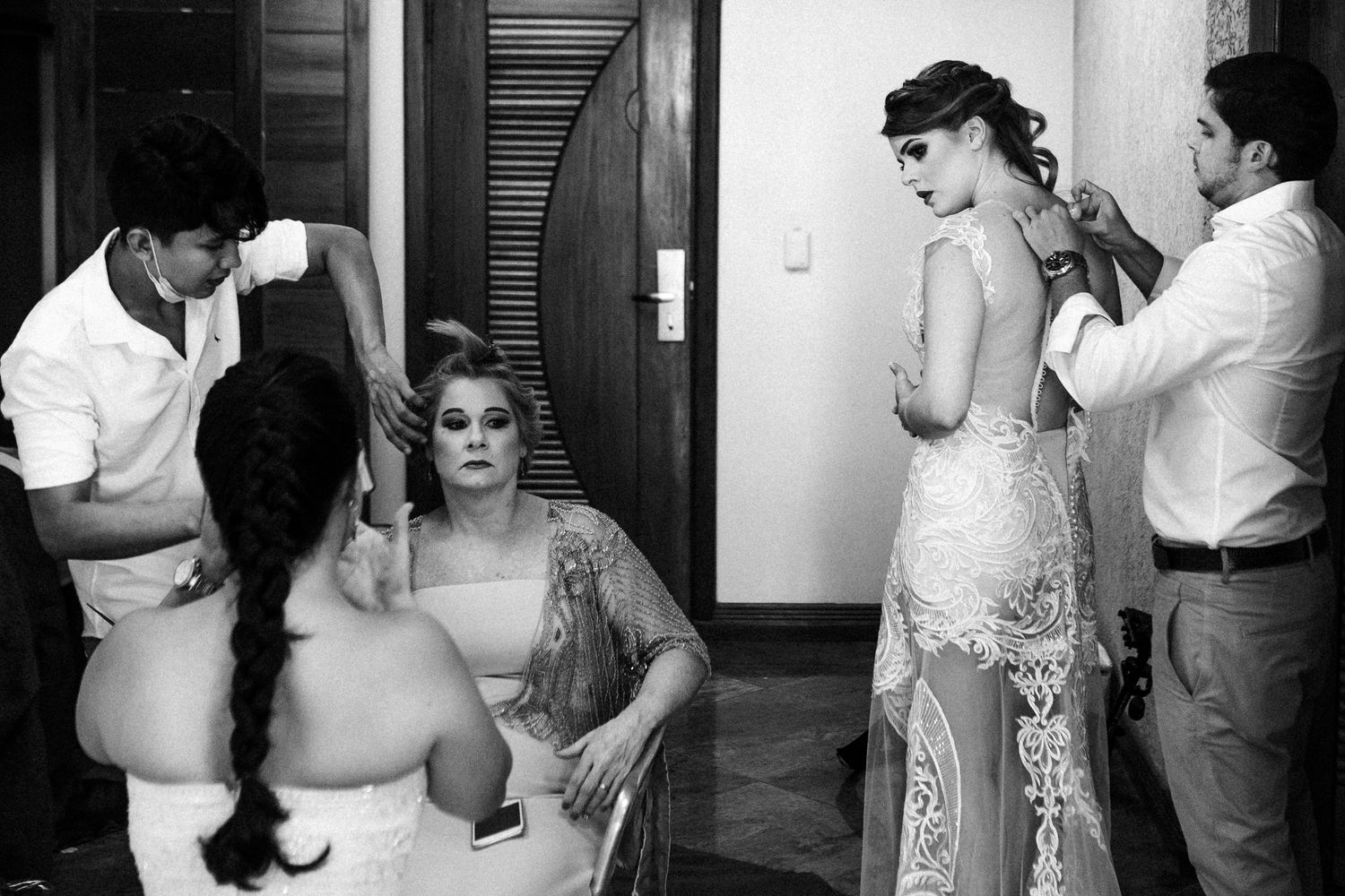 A black and white photo showing wedding preparation scene with stylists working on hair and makeup in a dressing room.