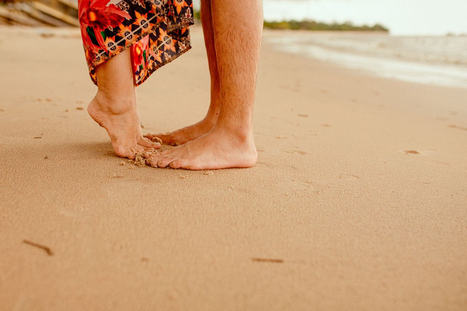 Two pairs of bare feet stand close together on a sandy beach with someone wearing red patterned clothing.