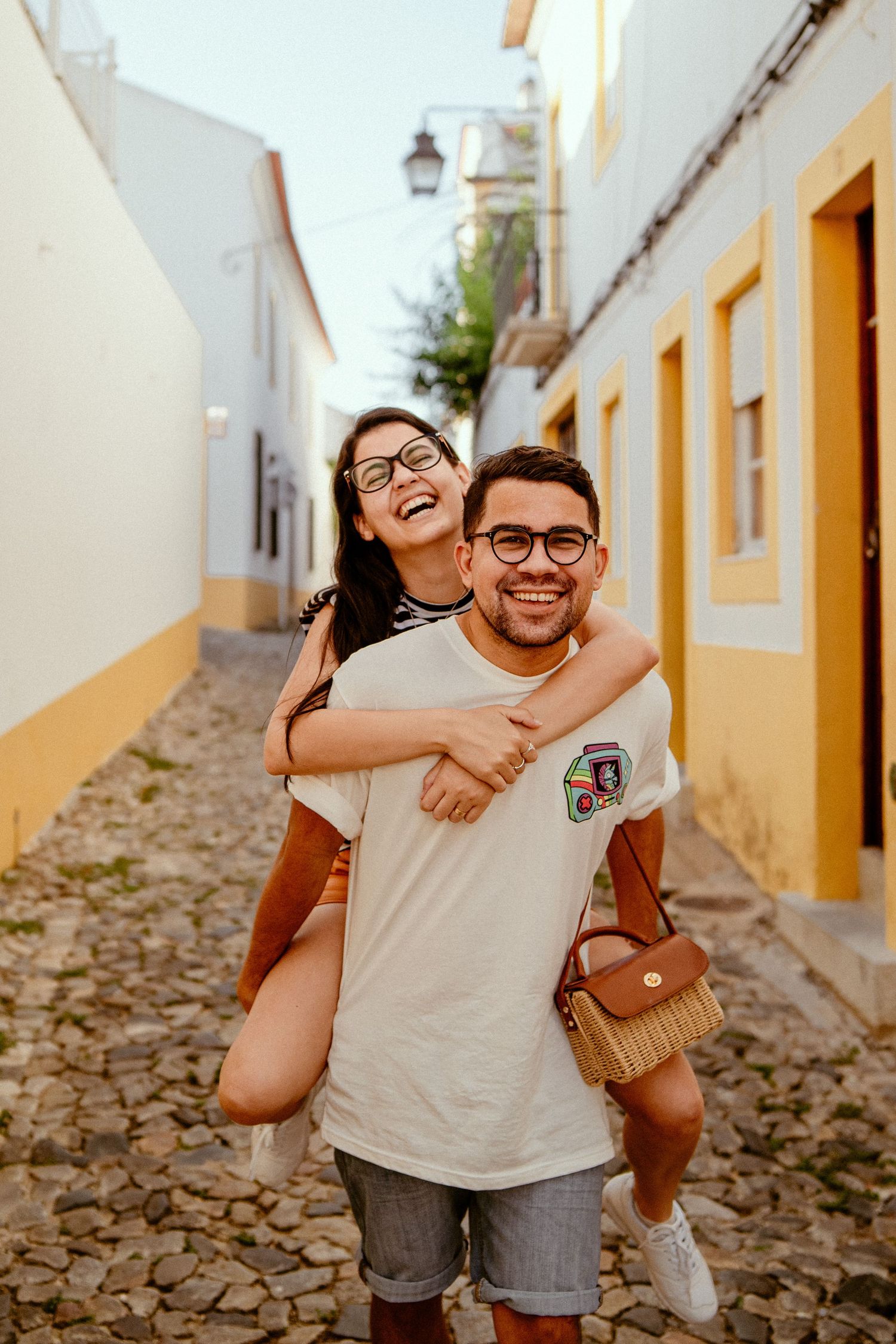A joyful couple sharing a piggyback ride moment on a charming cobblestone street with yellow and white buildings.