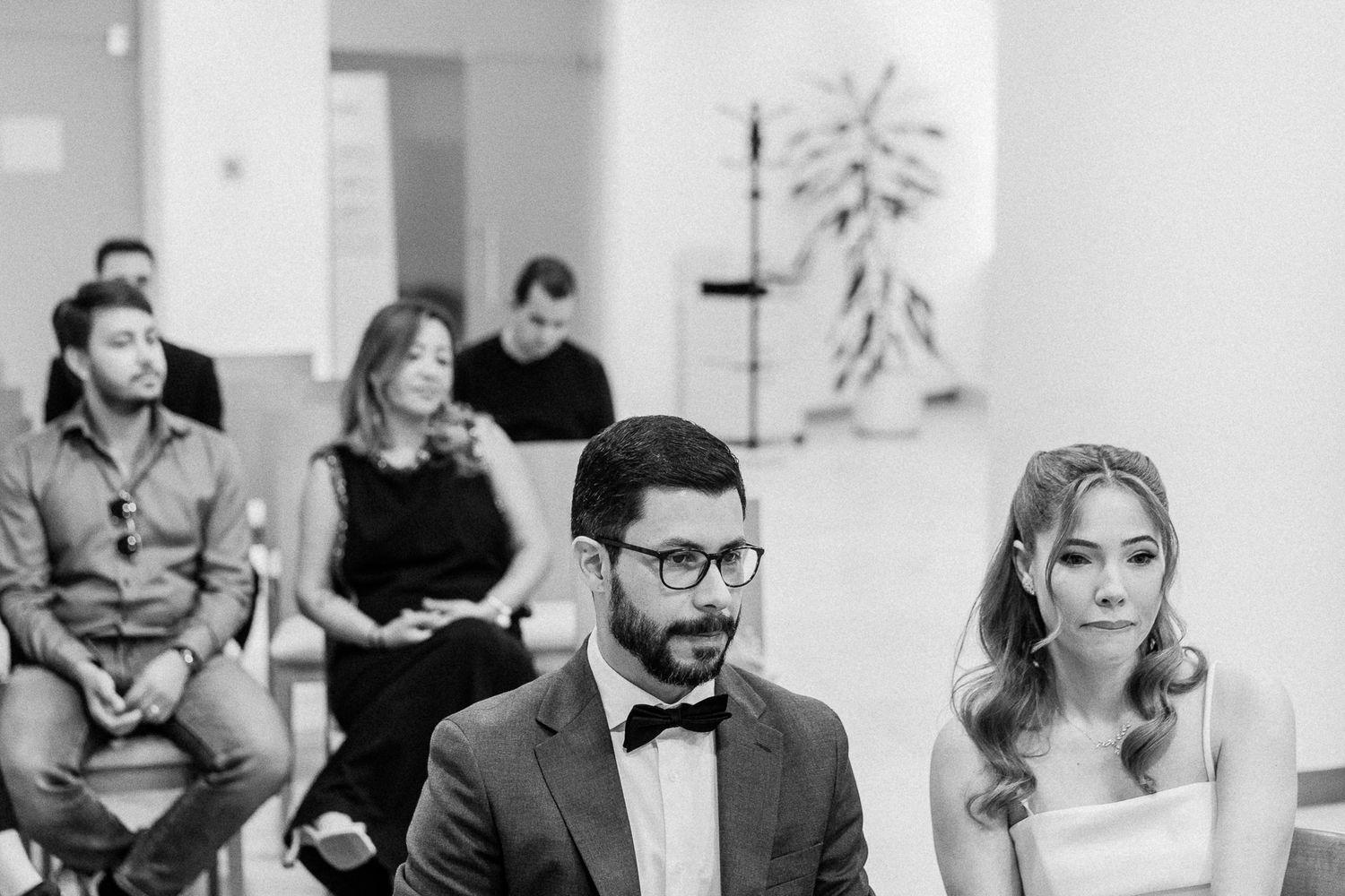 Bride and groom sit together at wedding ceremony with guests seated behind them in black and white photo.