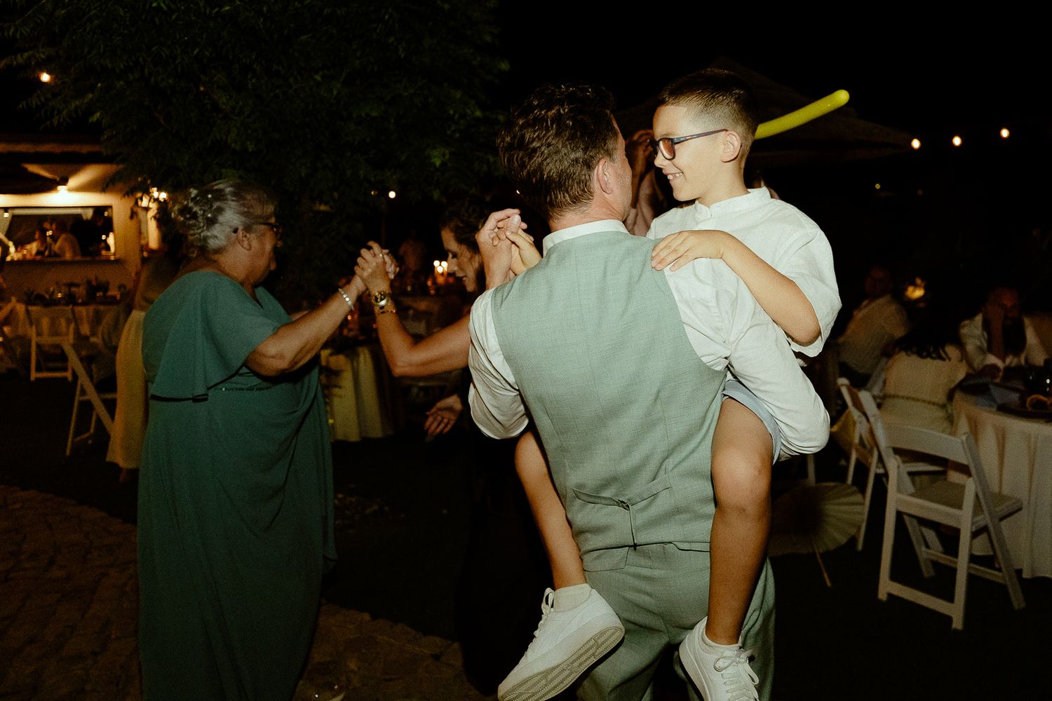 Two men dancing joyfully at an outdoor wedding reception at night, one lifting the other, with guests in background.
