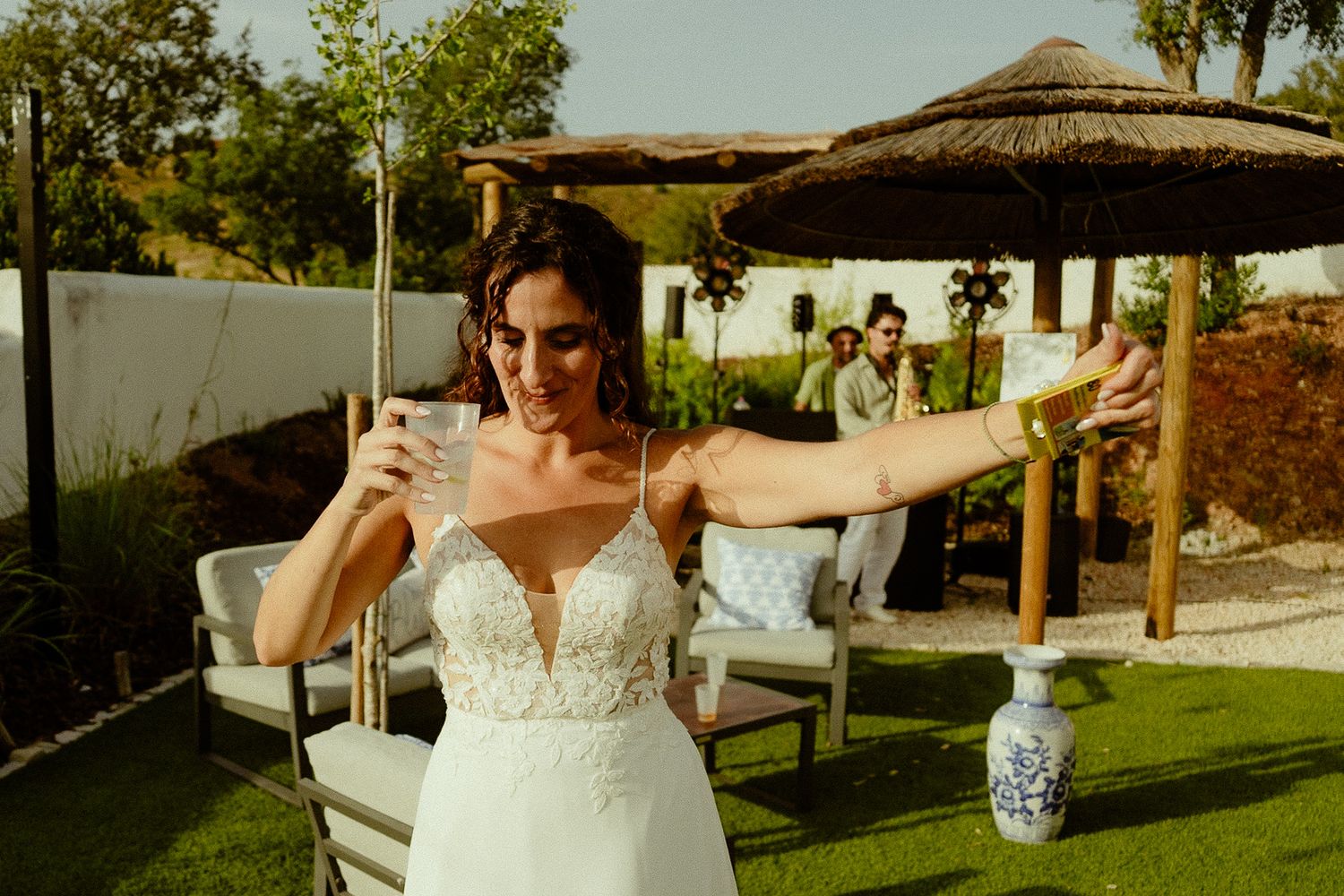 Bride in white lace gown holding a drink outdoors at a garden wedding venue with thatched umbrellas.