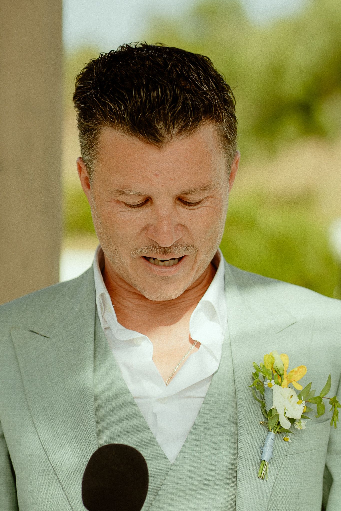 Groom in light green suit with yellow boutonniere reads vows at outdoor wedding ceremony.
