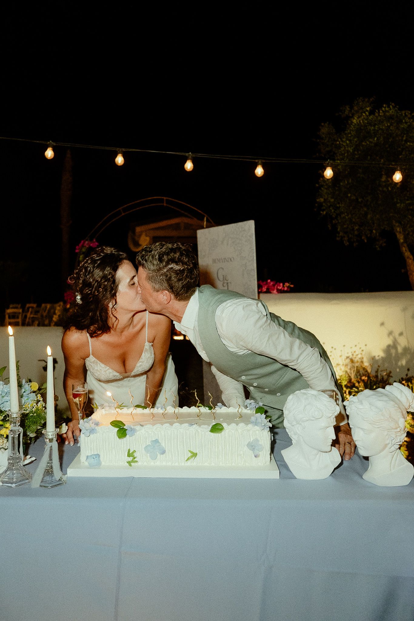Bride and groom sharing a kiss behind their white wedding cake at an outdoor evening reception.