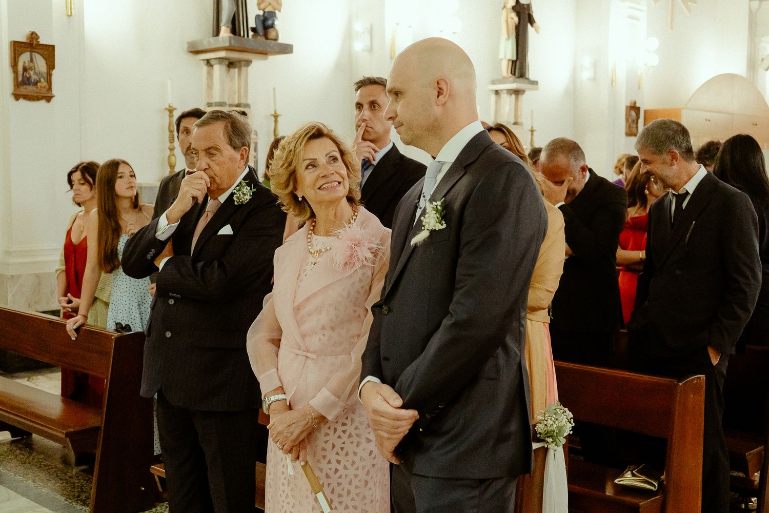 Guests stand in a church aisle during a wedding ceremony, dressed in formal attire, with pews and decor visible.