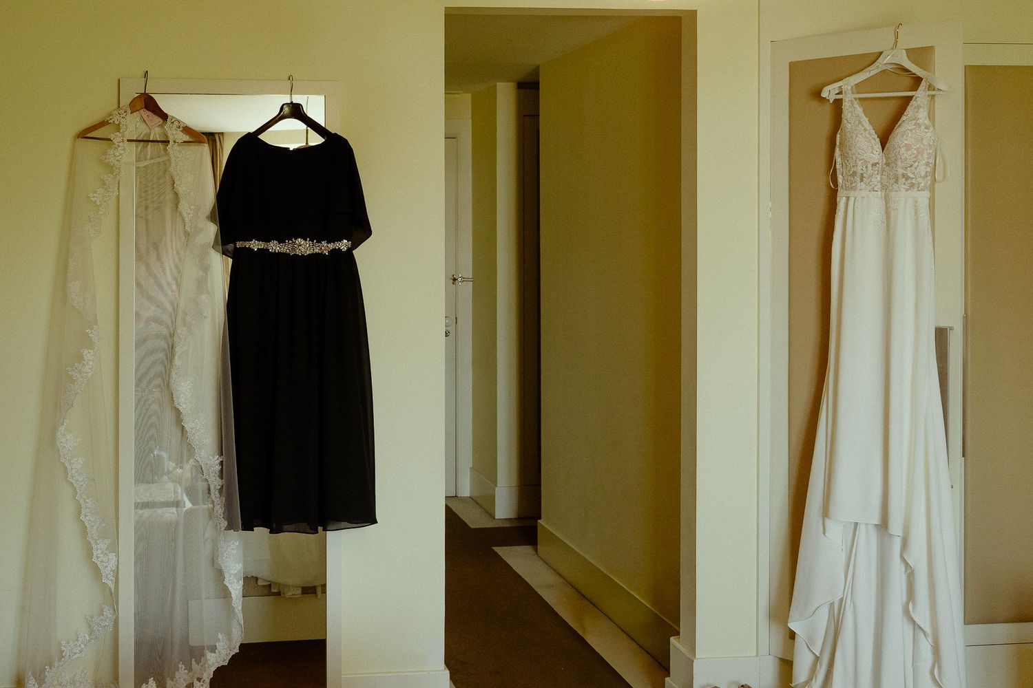 Three wedding dresses hanging in a bright room, including a white bridal gown, black bridesmaid dress, and ivory gown.